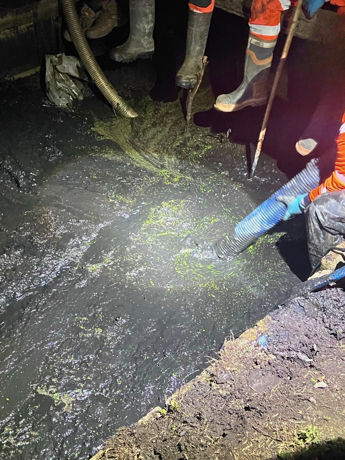 Workers wearing boots and gloves using hoses and tools to manage a thick, dark, and greenish liquid or sludge in a confined space.