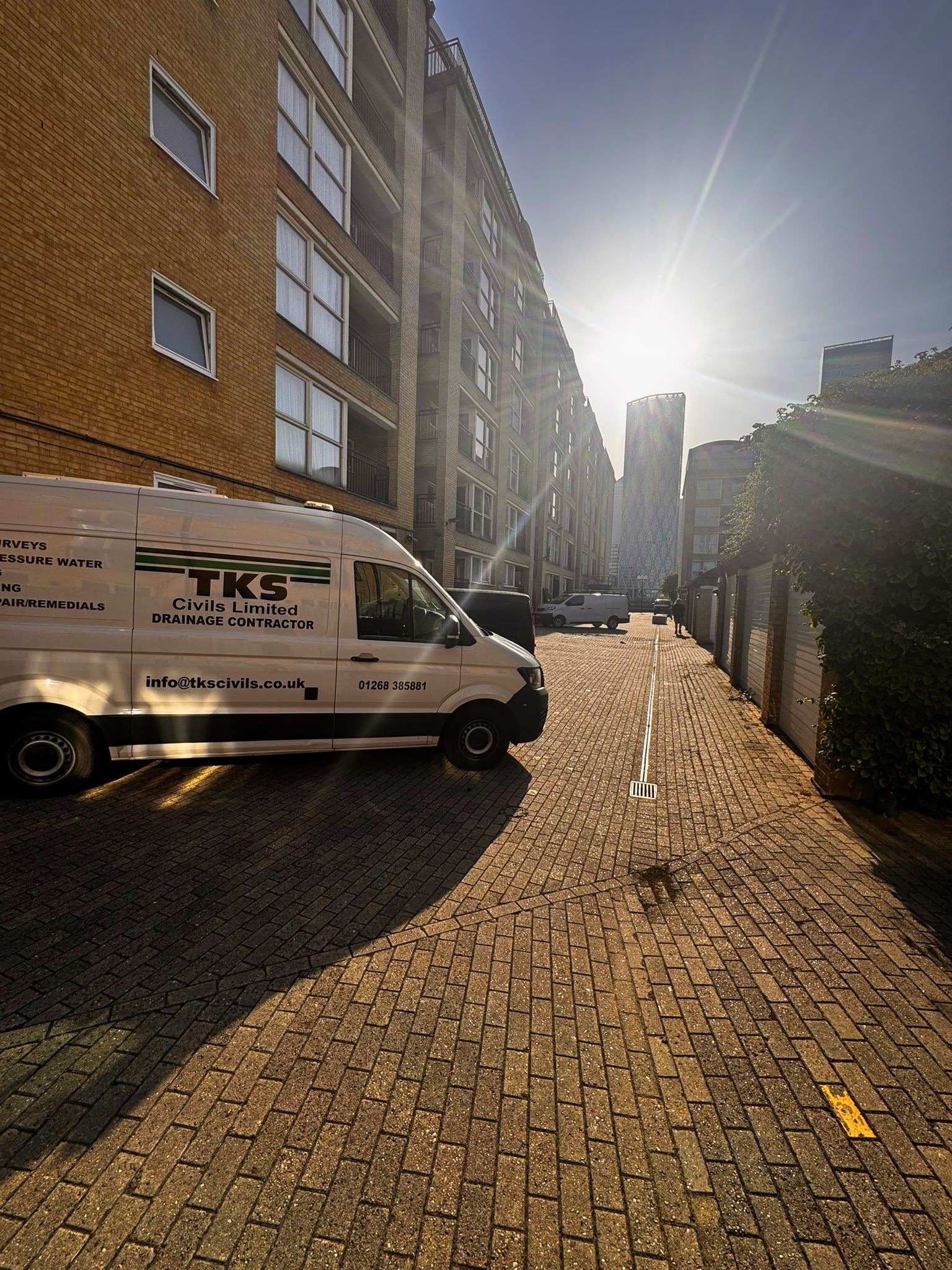 A city street scene during daytime featuring a white work van with black and green text that reads 'TKS Civil Limited Drainage Contractor' parked along a brick-paved sidewalk. The buildings are modern, with multiple floors, balconies, and large windows, with taller skyscrapers visible in the background. The sun is shining brightly, causing a lens flare and casting shadows on the street.