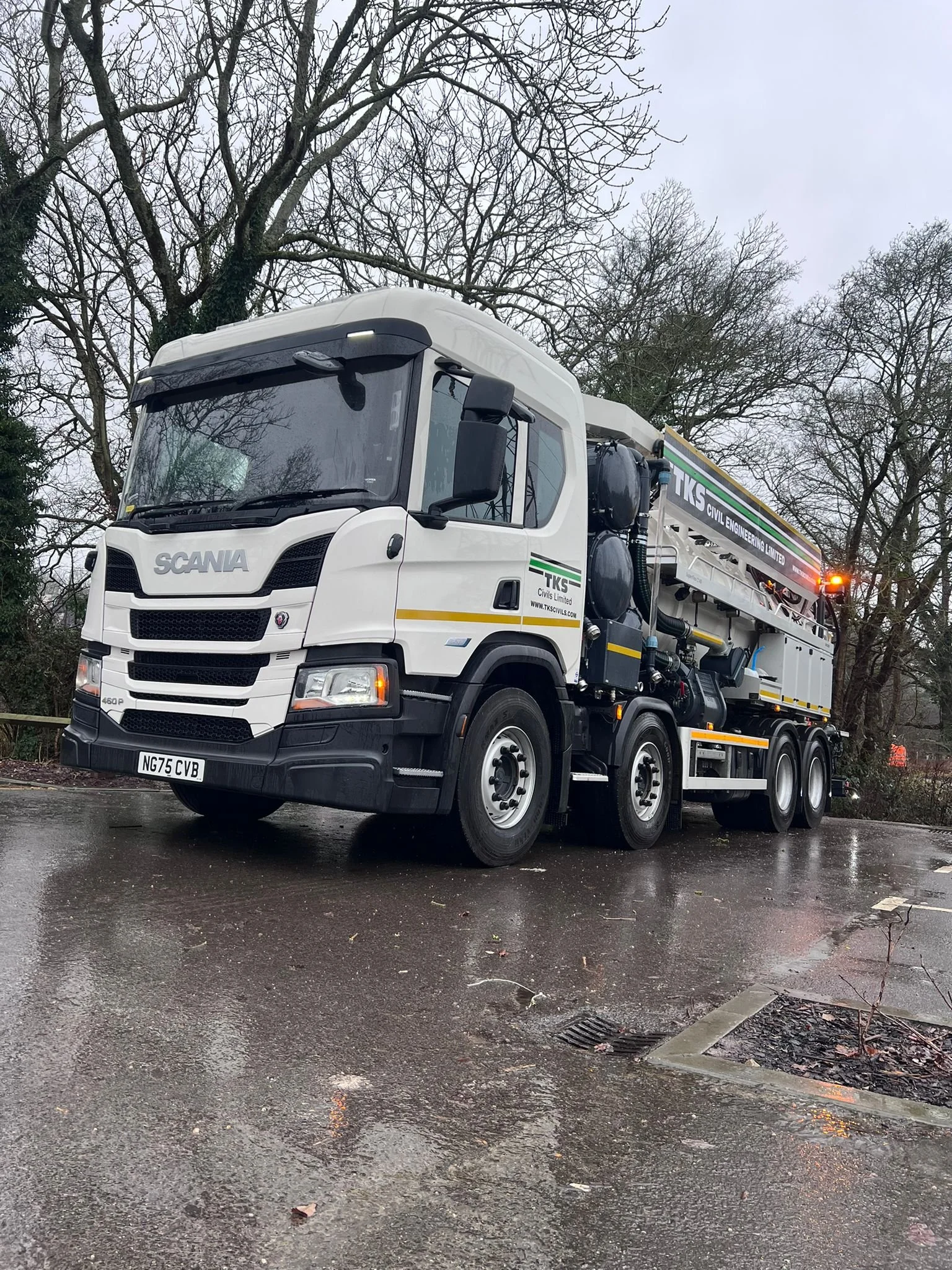 A white industrial truck parked on a wet pavement on a cloudy day, with trees without leaves in the background.