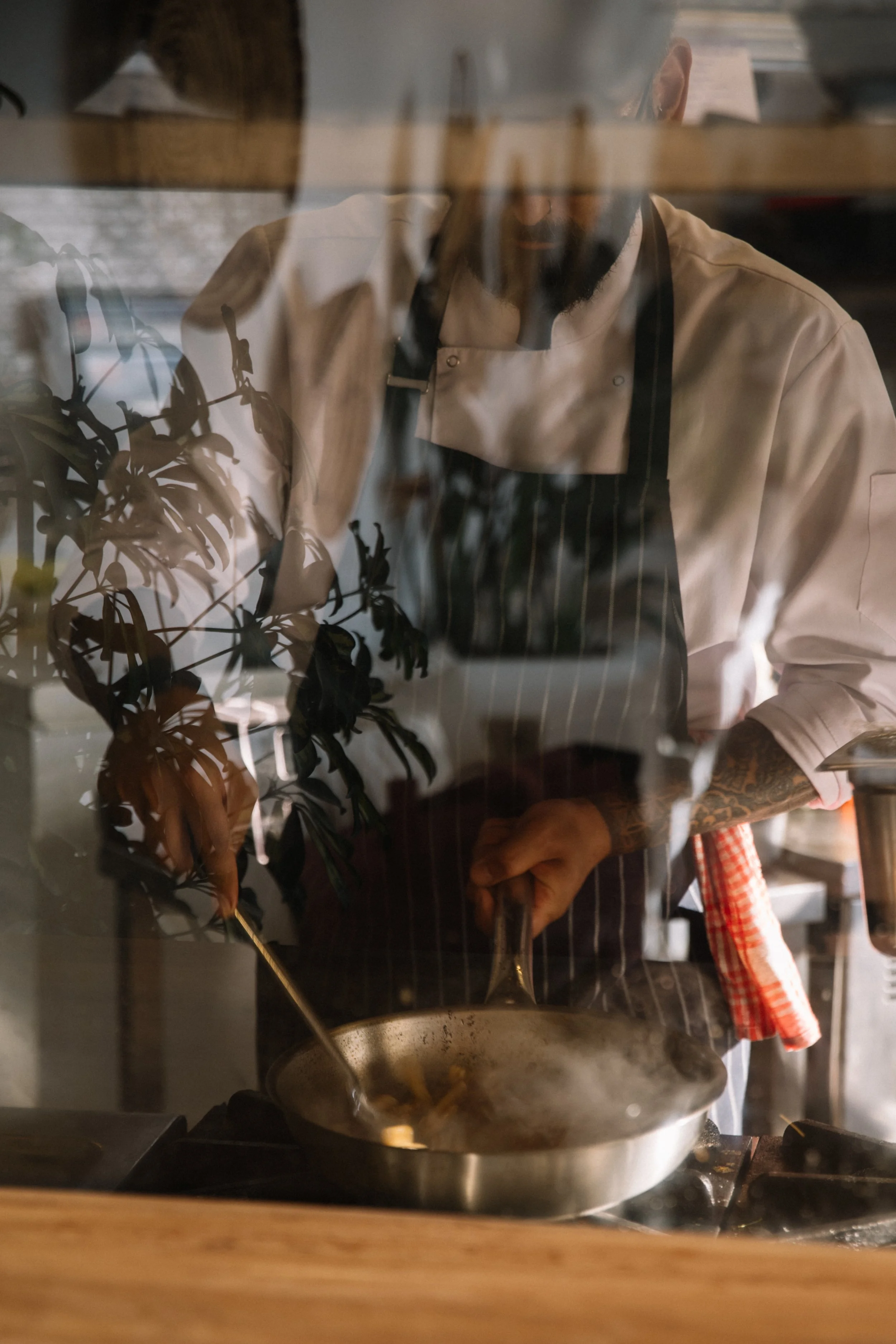 A chef with a beard and tattoos wearing a beige chef's jacket and black apron cooking in a kitchen, viewed through a glass window with reflections of plants and outdoor scenery.
