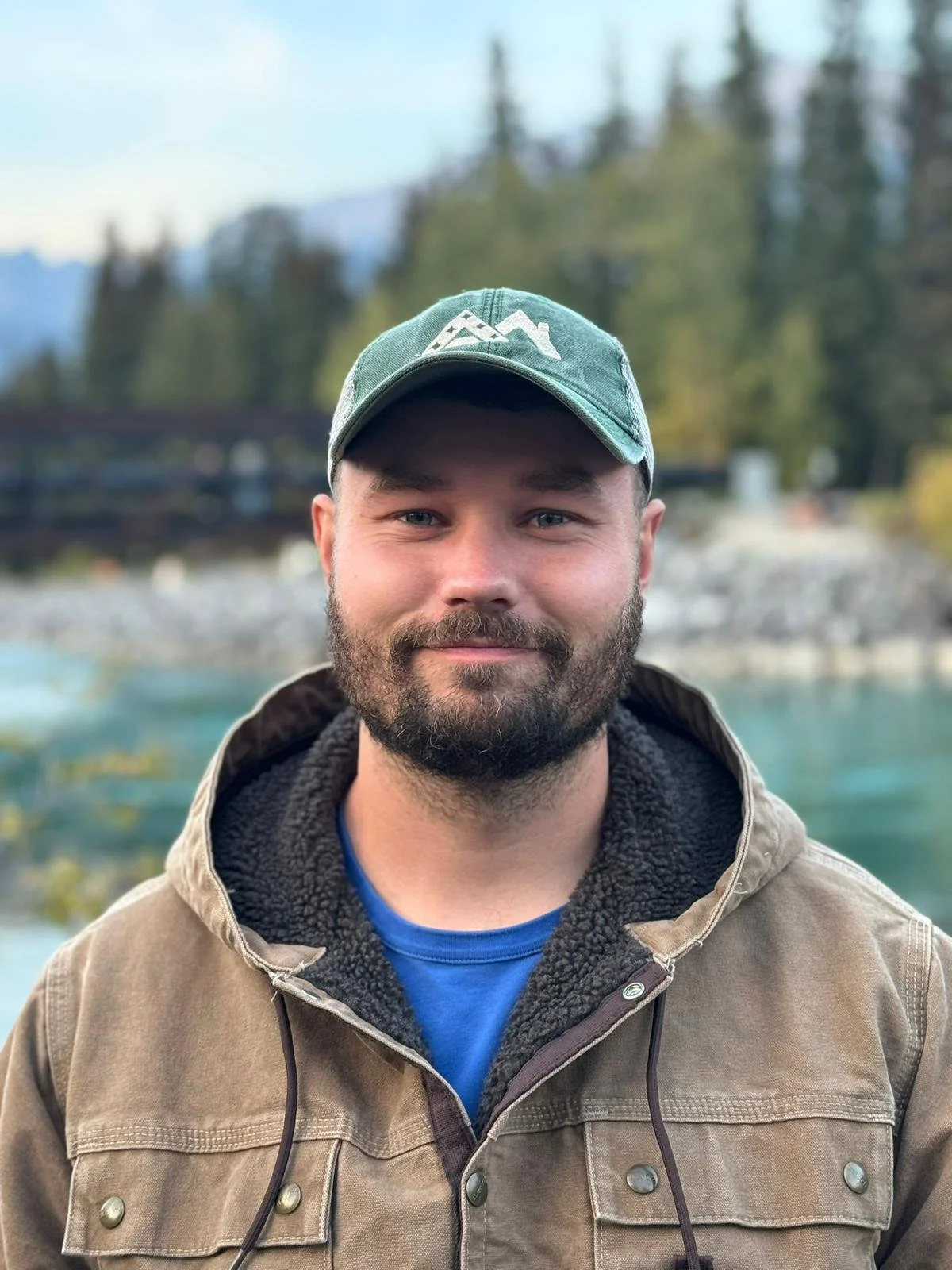 A man with a beard and blue eyes wearing a green cap, brown jacket, and blue shirt outdoors near a body of water with trees and mountains in the background.