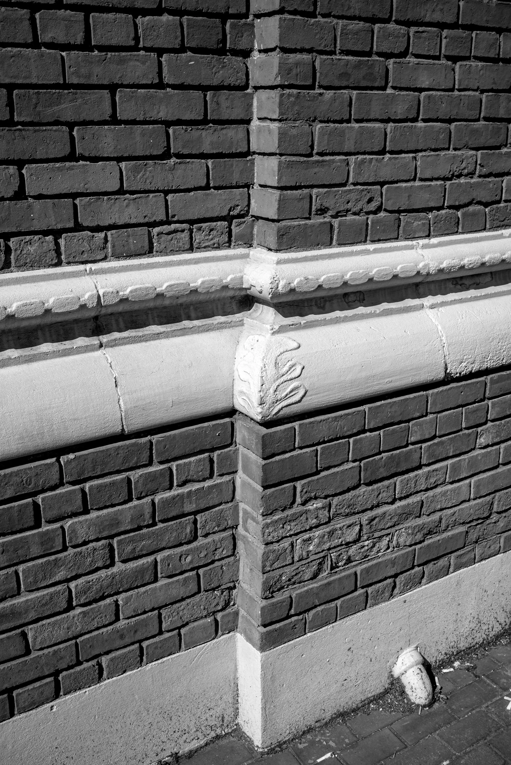 Close-up of a brick building corner with decorative stonework and a drain pipe on the ground.