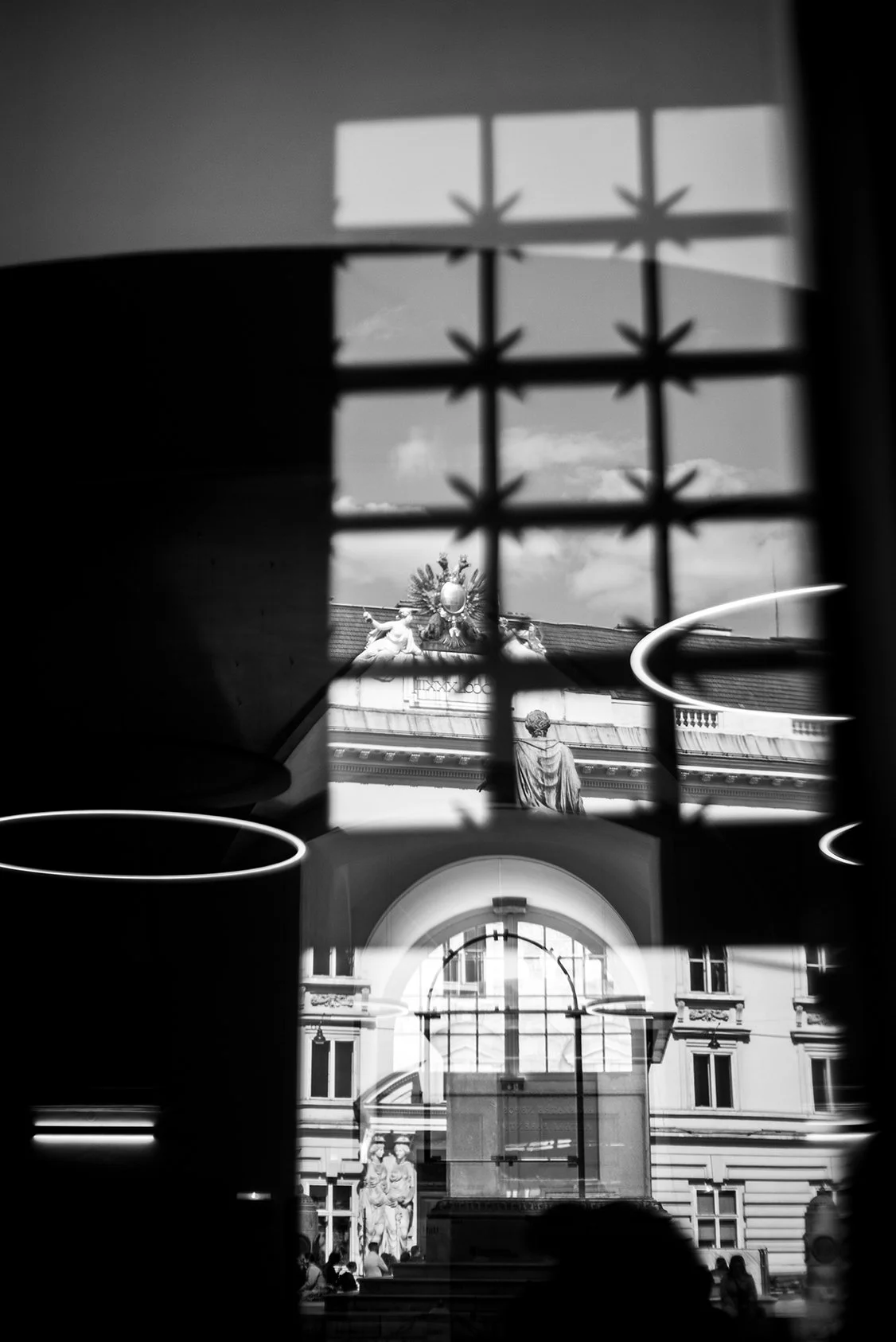 A black and white photograph showing an outdoor scene through a window with a decorative metal grille. The scene includes a building with ornate sculptures and statues, a large arched window, and several clouds in the sky.