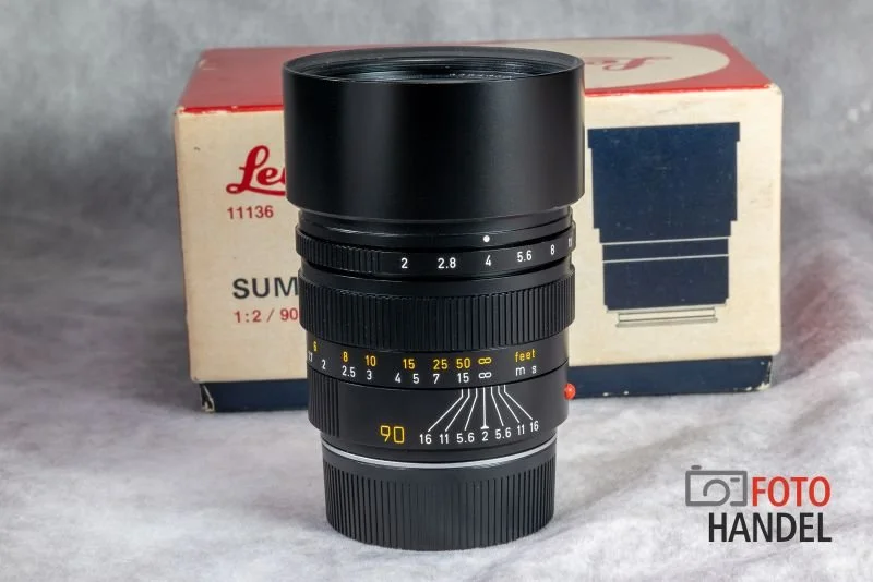 A vintage camera lens with a black exterior, multiple adjustable rings with markings for aperture, focus distance, and depth of field, positioned in front of its original box with red and white colors.