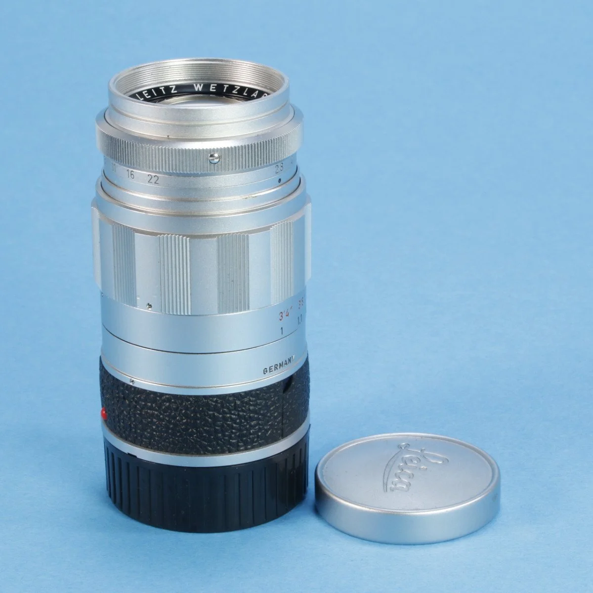 A vintage silver camera lens with a black textured grip, placed against a light blue background, with a silver lens cap beside it.