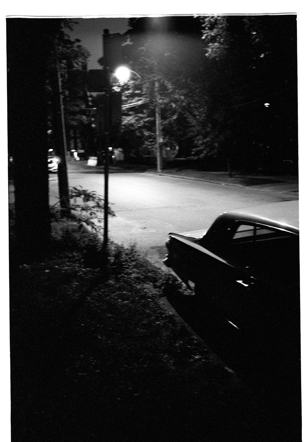 A black and white photo of a quiet street at night, showing a vintage car parked on the side, a streetlamp illuminating the scene, and trees lining the road.