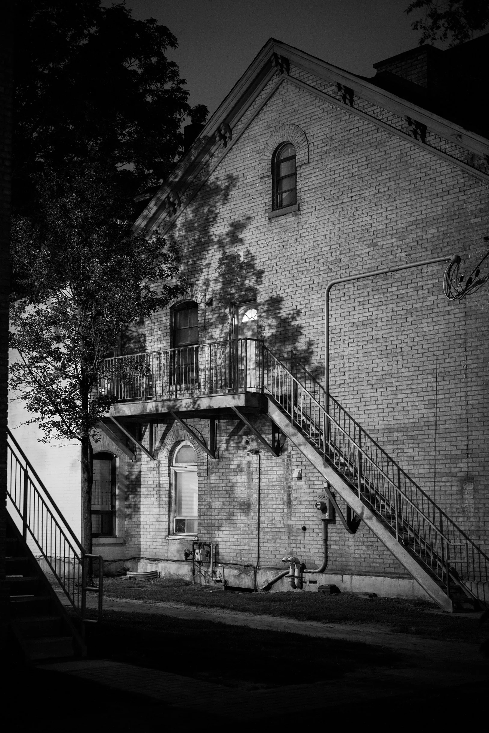 Black and white photo of the side of a brick building at night, with a staircase leading up to a second-floor balcony, a tree casting shadows on the building, and trees visible at the top of the image.