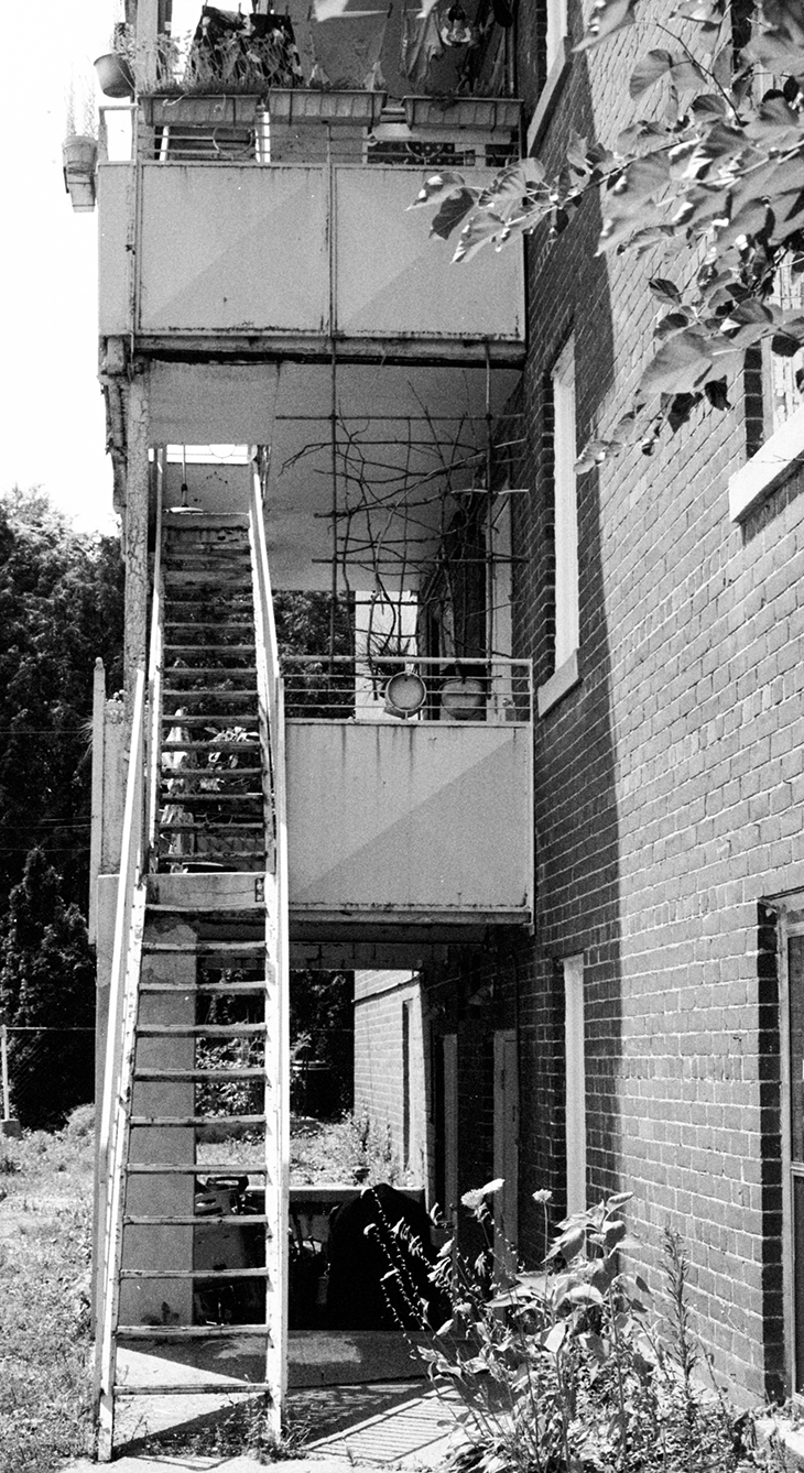 An outdoor metal staircase leading to a second-floor balcony of a brick building with some hanging plants and outdoor items on the balcony.