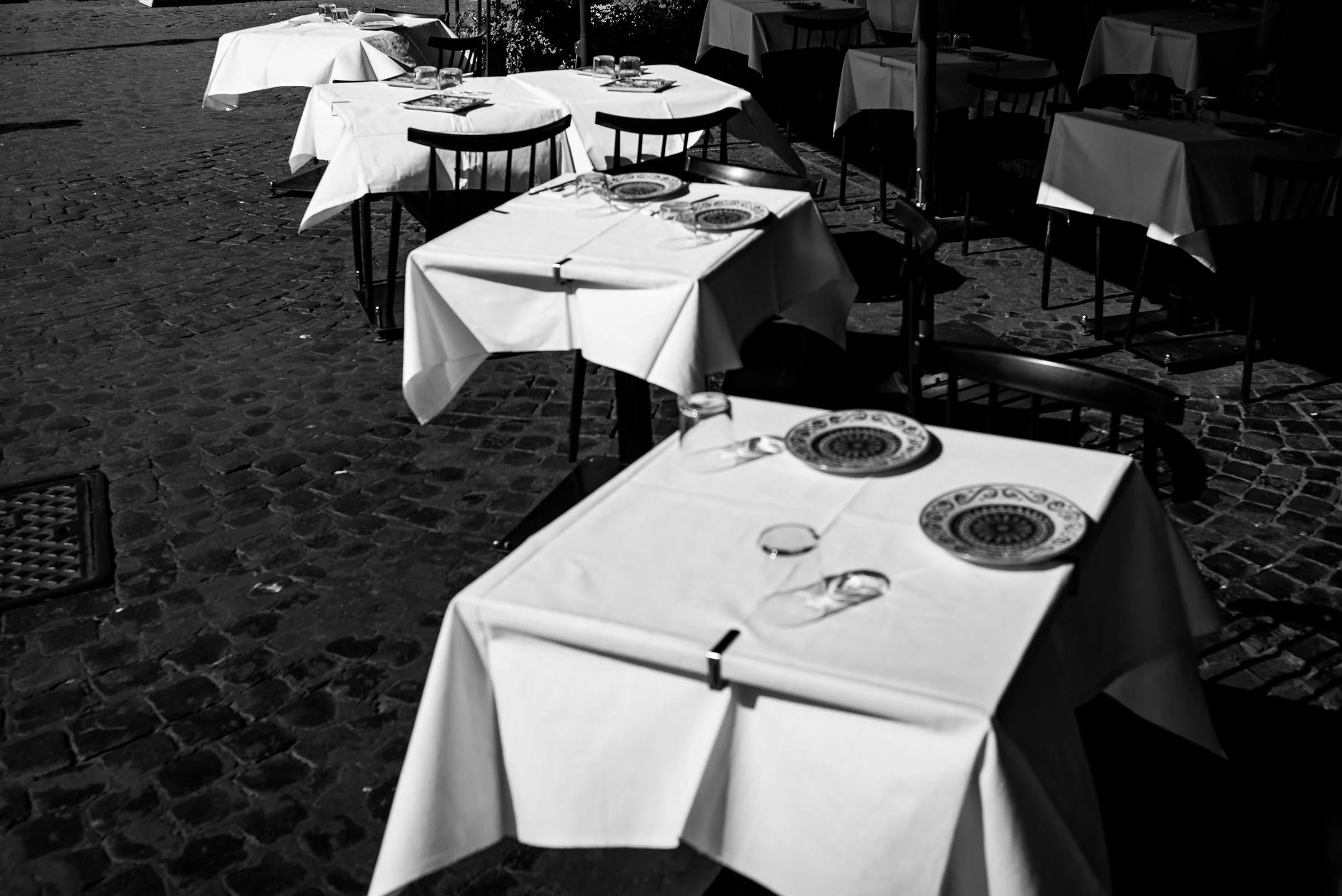 Empty outdoor restaurant patio with tables covered in white tablecloths, set with plates, glasses, and utensils on a cobblestone ground.