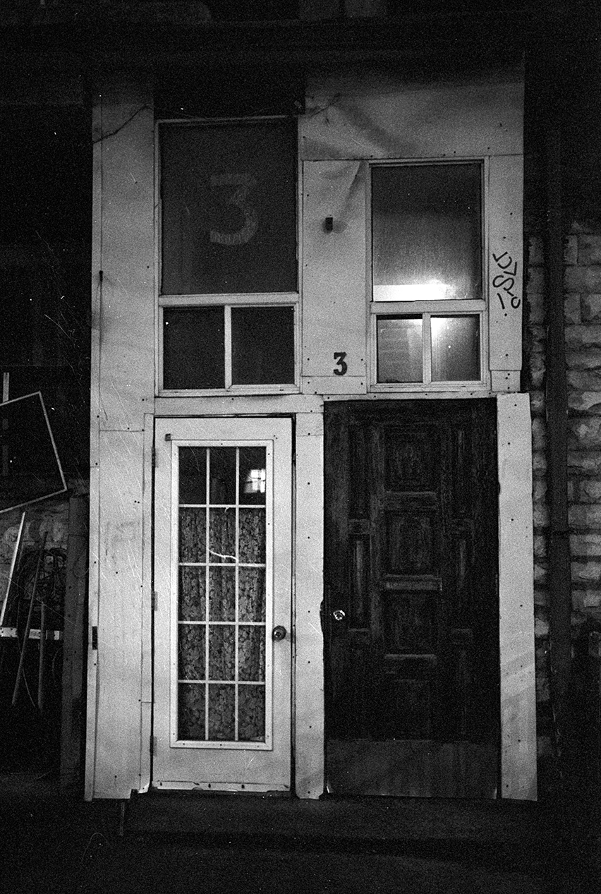 Black and white photograph of a building's front entrance with a door and three windows above it. The door on the left has glass panes, and the door on the right appears to be solid wood. The number '3' is painted on the wall next to the windows and on the window glass.