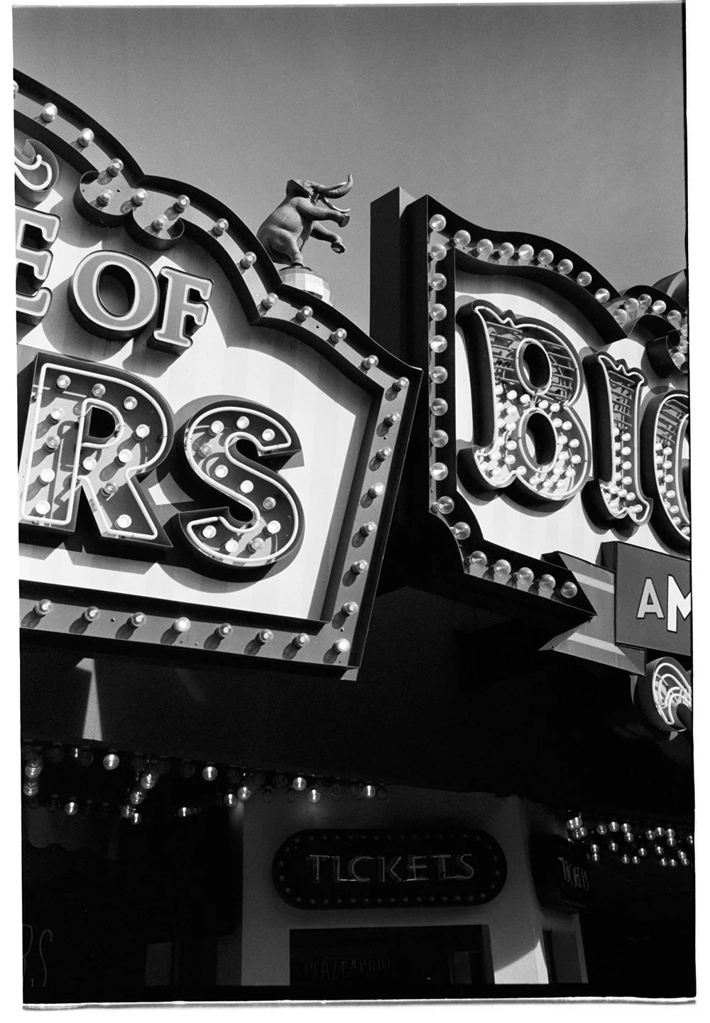 Black and white photo of a carnival or amusement park marquee with illuminated bulbs, featuring the words "TICKETS" and part of a sign with the word "BEARS," and a small statue of a bison or buffalo on top of the marquee.