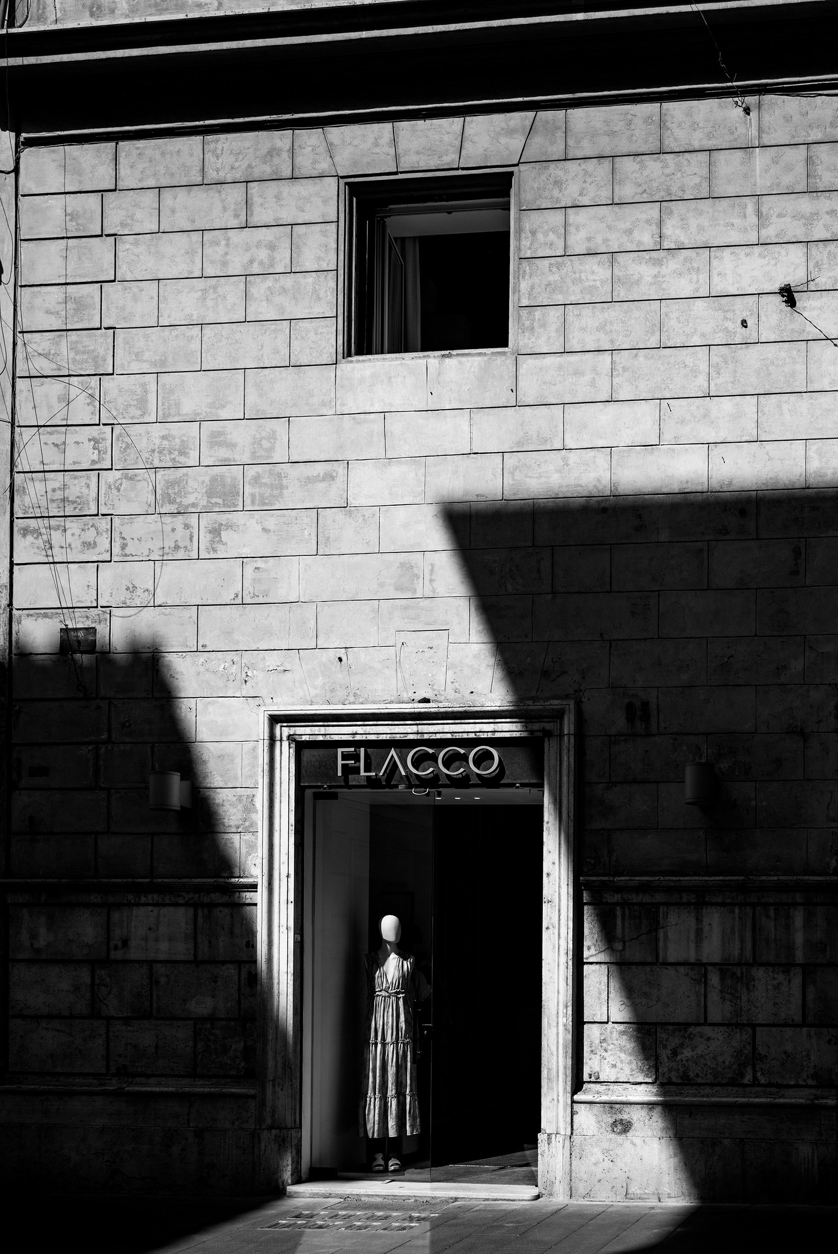 Black and white photo of a building's facade with a store called Flacco. The store's entrance has a mannequin wearing a dress in the doorway. There is a window above the store, and shadows cast across the stone wall.