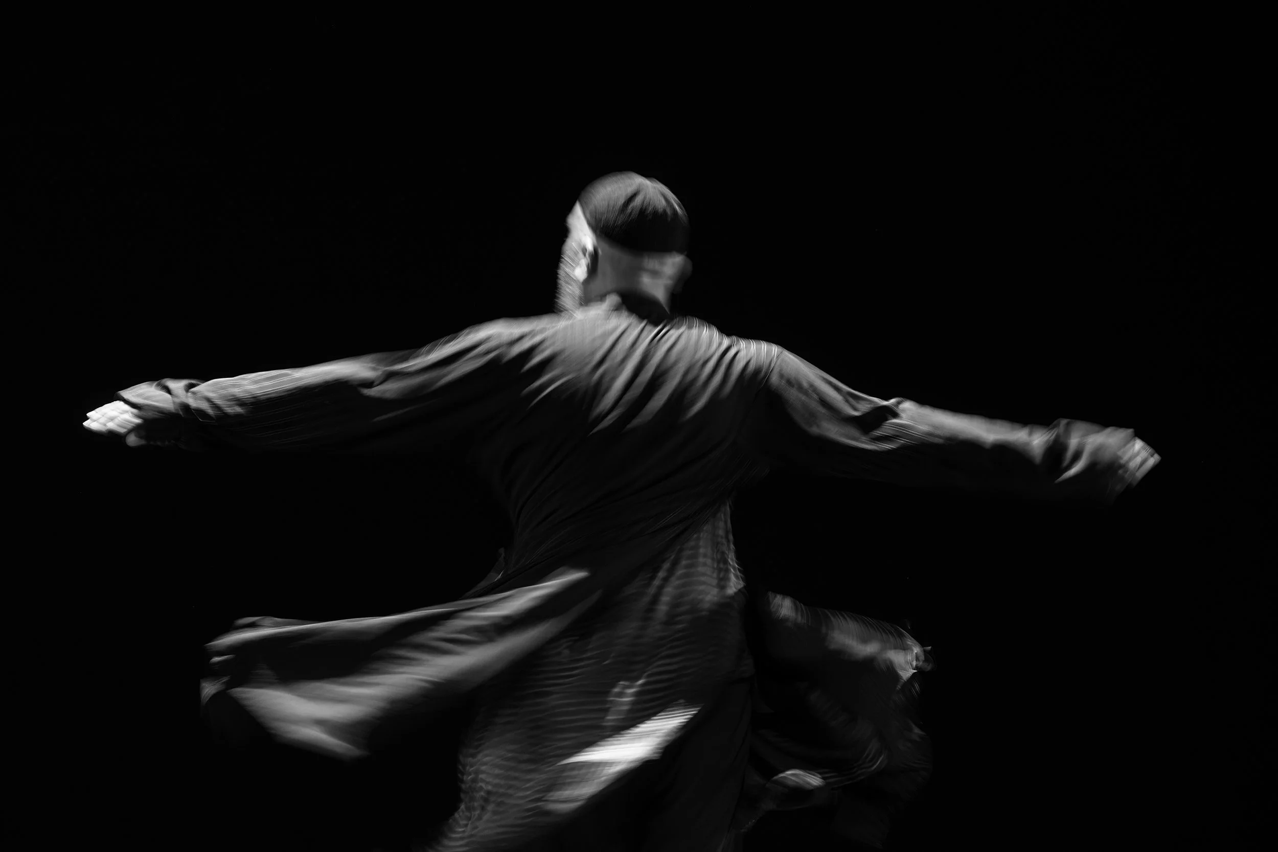 A person spinning or dancing with arms extended, wearing flowing clothing, against a black background.