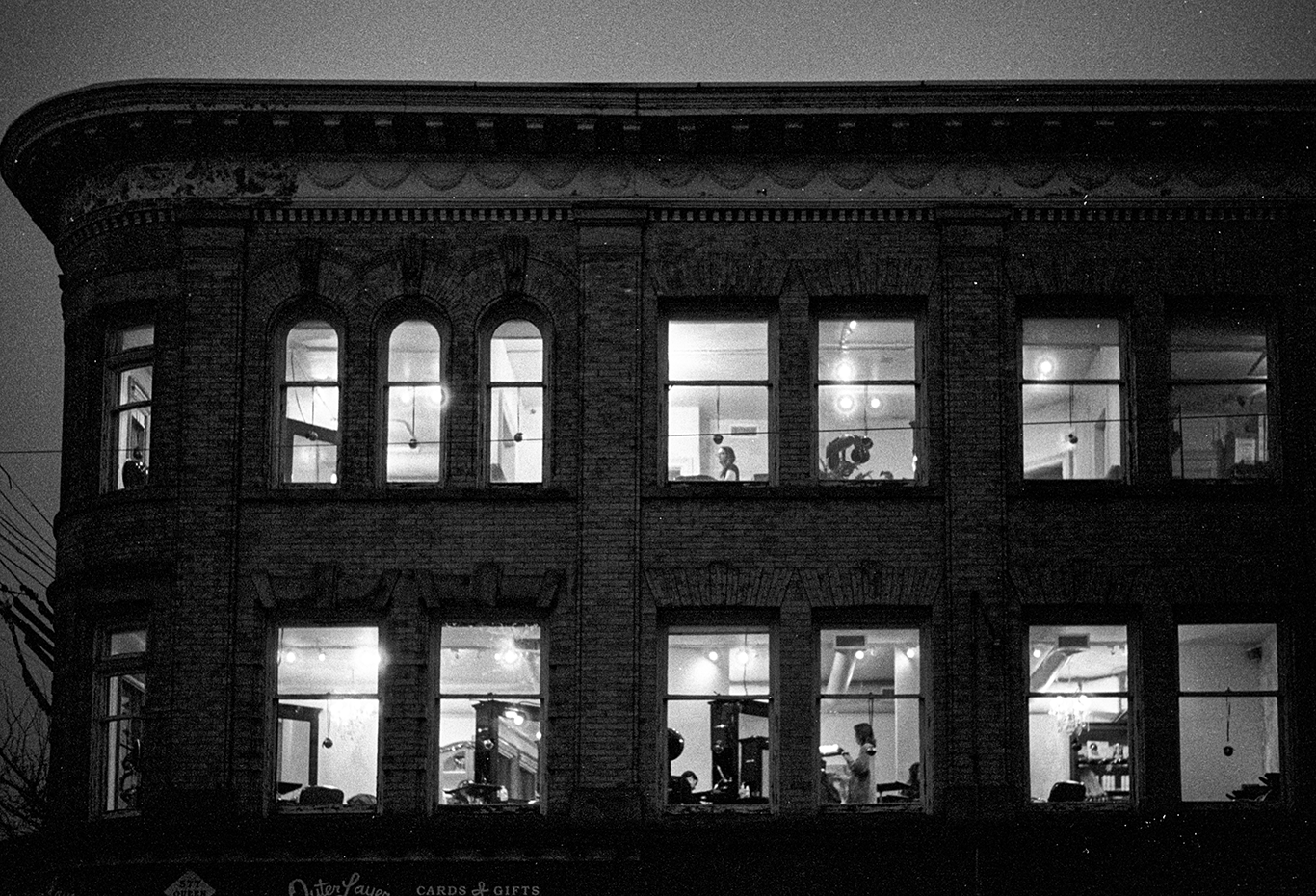 Nighttime view of a three-story brick building with illuminated interior seen through large windows, showing people working or exercising inside.