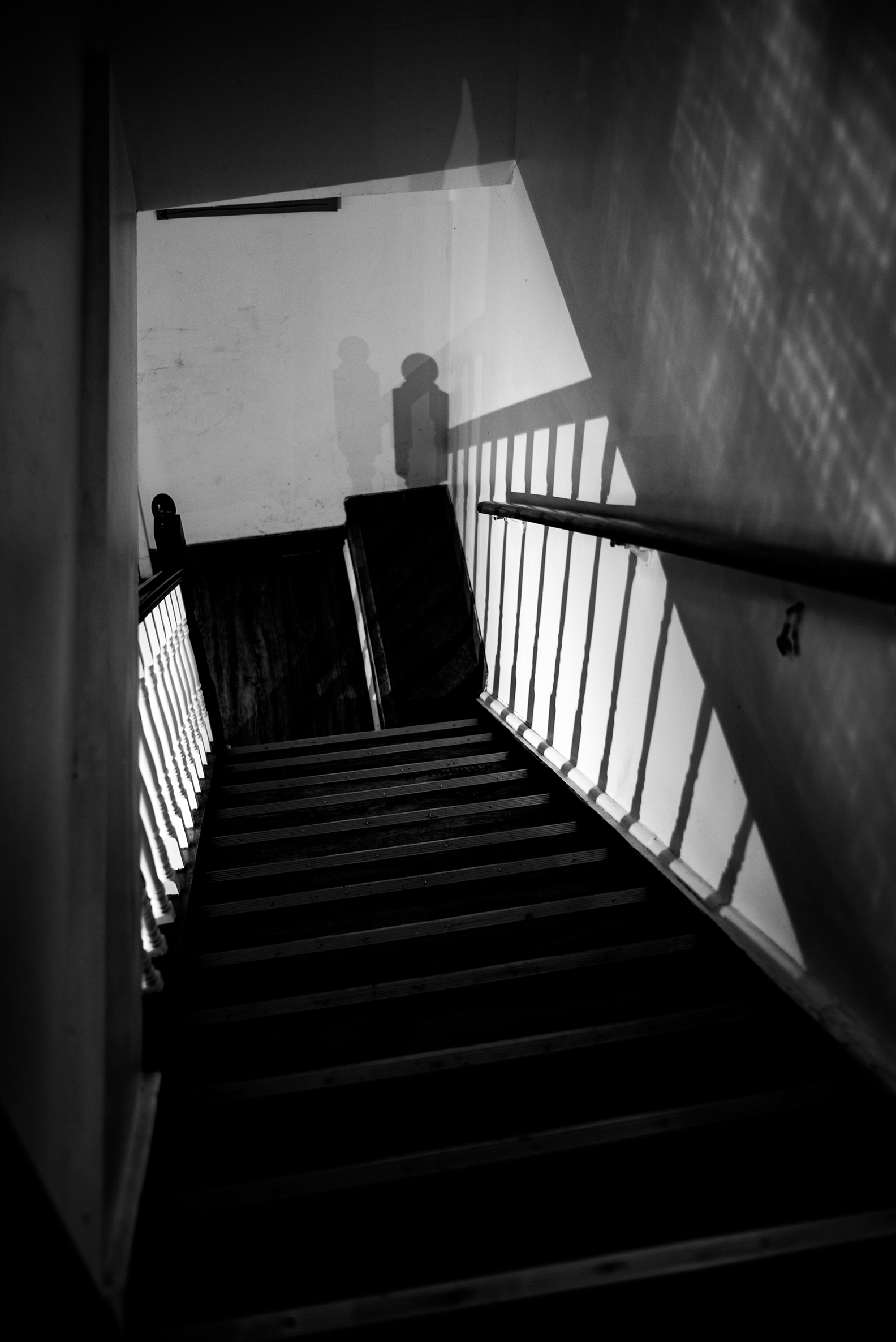 Black and white photo of a staircase viewed from above, with shadows cast on the wall showing two people standing at the top of the stairs.