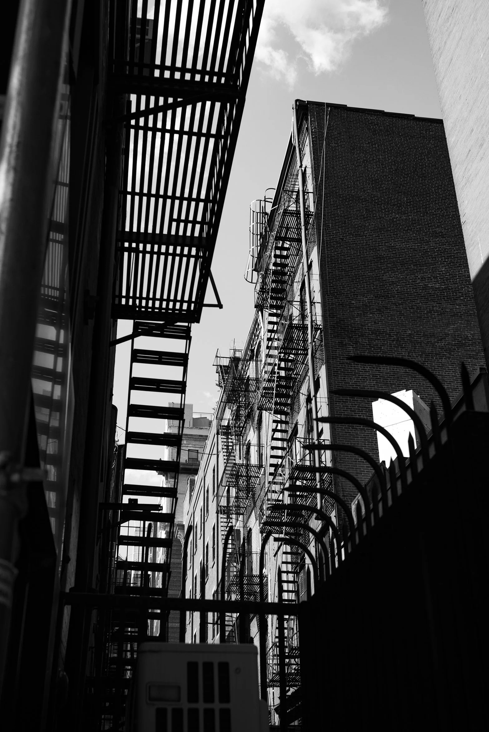 Black and white photo of fire escapes on urban buildings with metal staircases and railings, viewed from below.