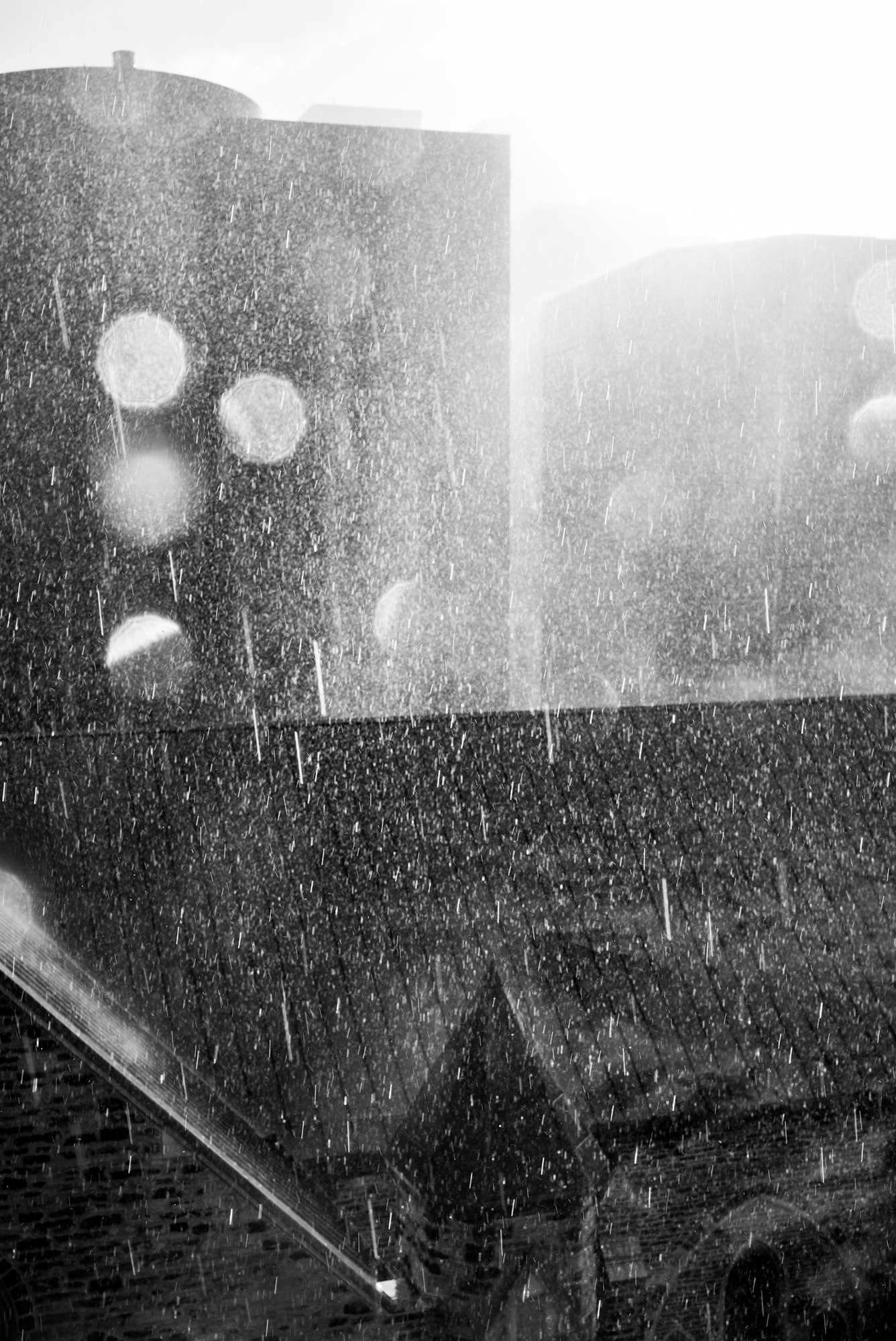Black and white photo of rain falling with city buildings in the background and a wet, shiny rooftop in the foreground.