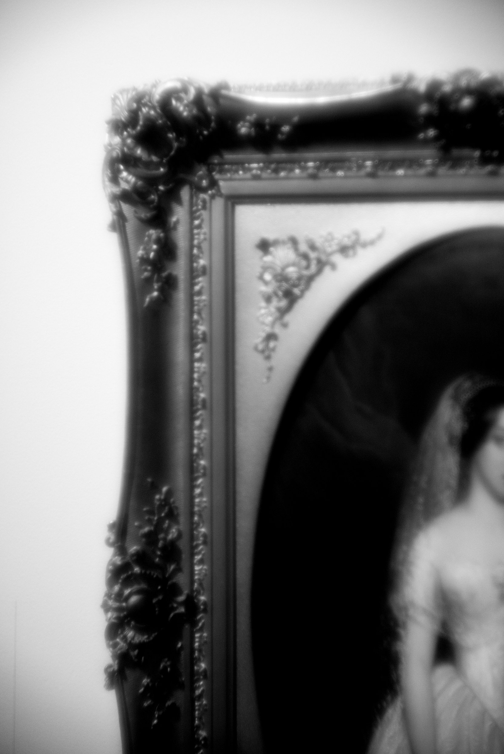 Part of an ornate picture frame with decorative floral and scroll details, with a mirror reflecting a person taking a photo, black and white photo.