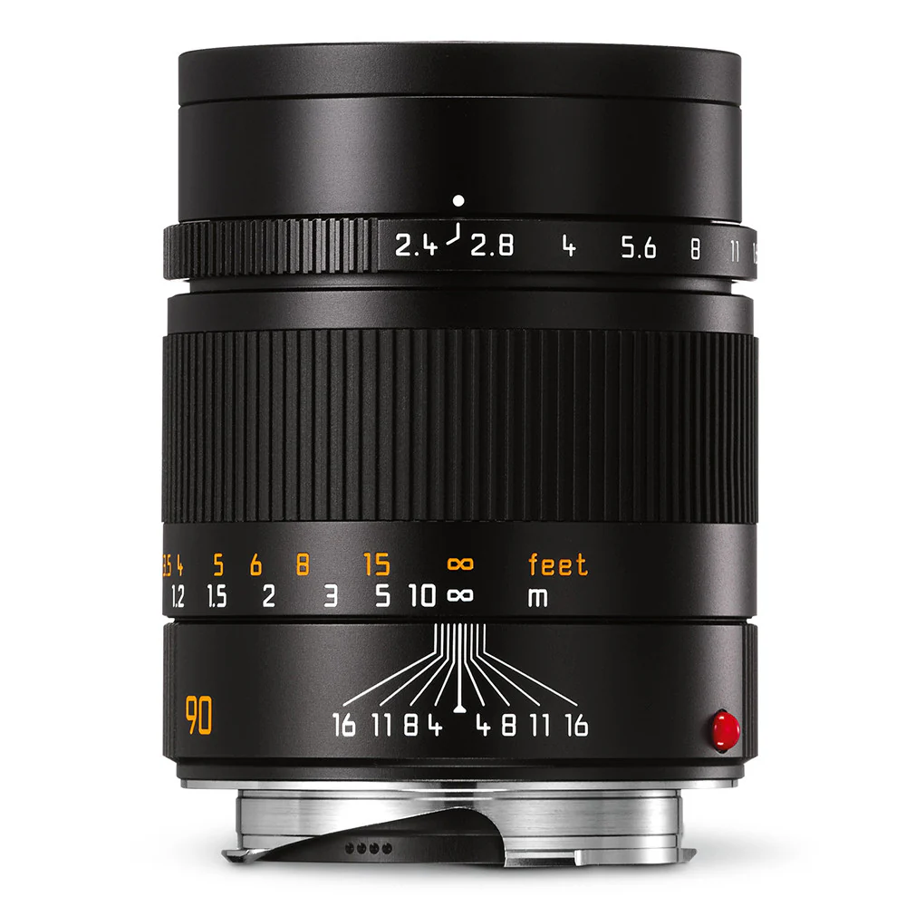 Close-up of a black camera lens with various focus and distance markings in white, orange, and yellow, and a red dot on the side.