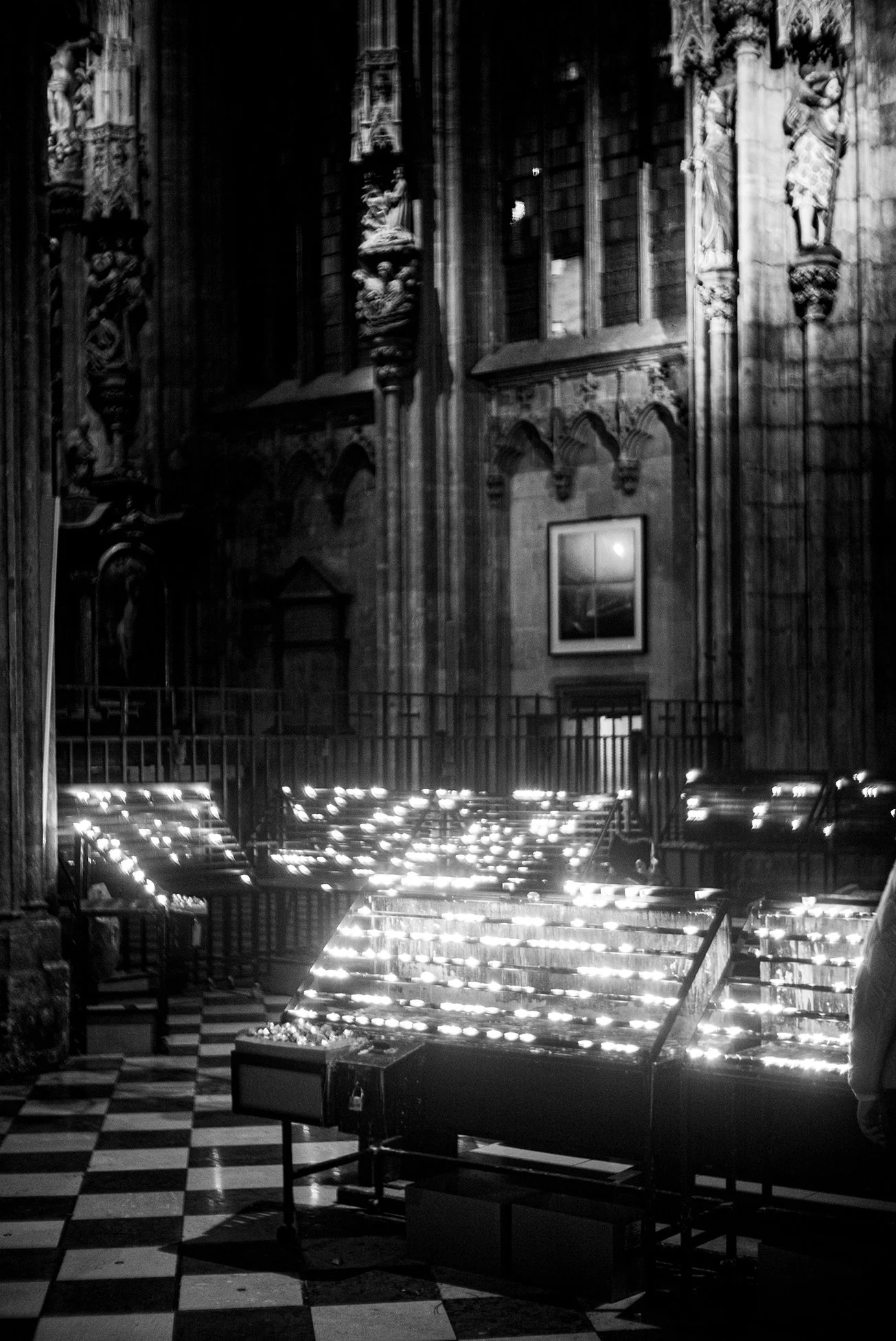 Black and white photo of the interior of a church with ornate stone carvings on the walls and stained glass windows. There are illuminated candle stands in the foreground and a checkered floor.