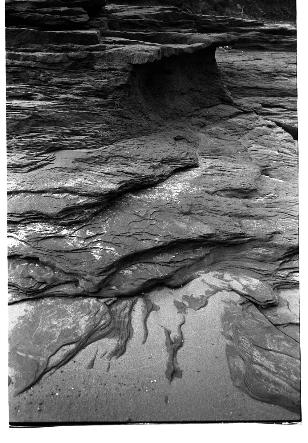 Black and white photo of layered rocks and sand at a beach with a small eroded cave or indentation.