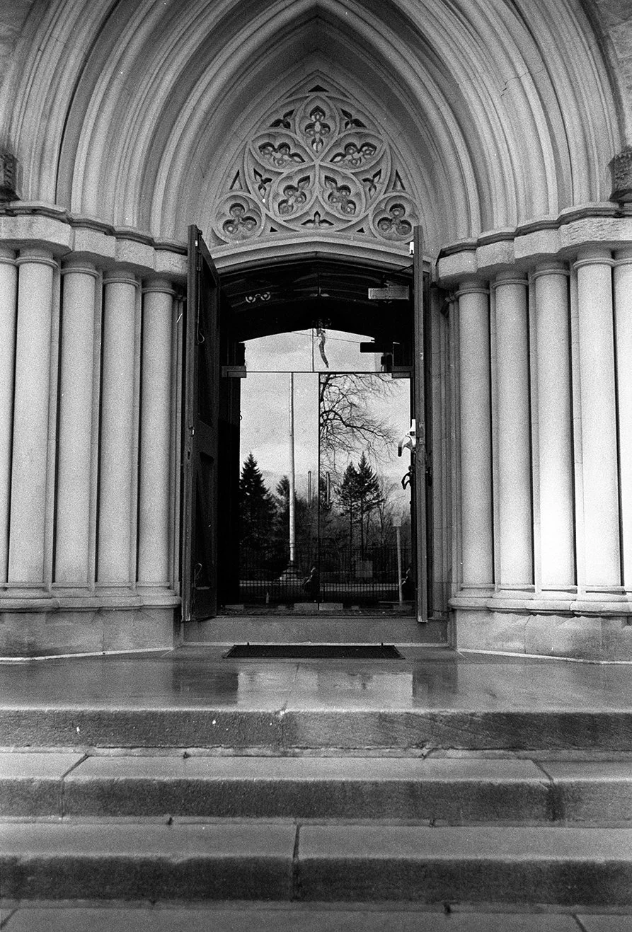 Black and white photo of the entrance to a church with stone columns and an ornate pointed arch above double glass doors that reflect trees and a cloudy sky.