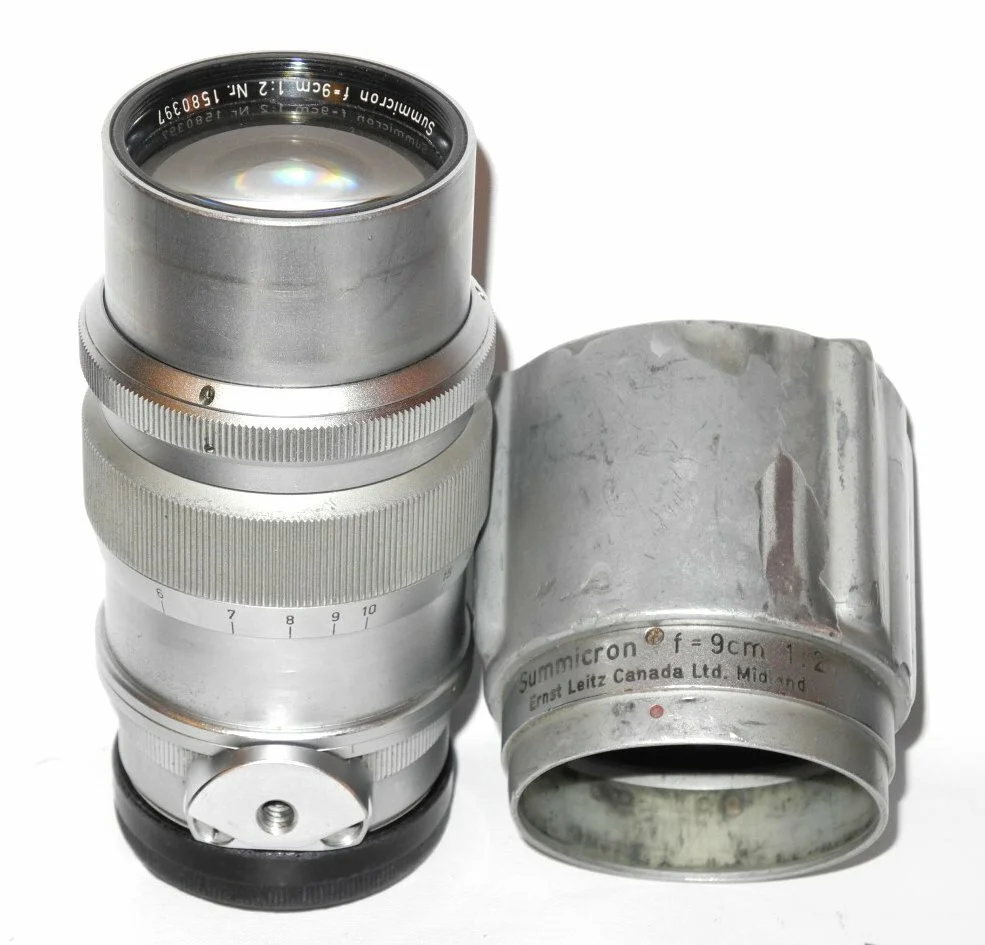 A vintage silver camera lens with an attached metal viewfinder lens, marked with manufacturer details, against a white background.