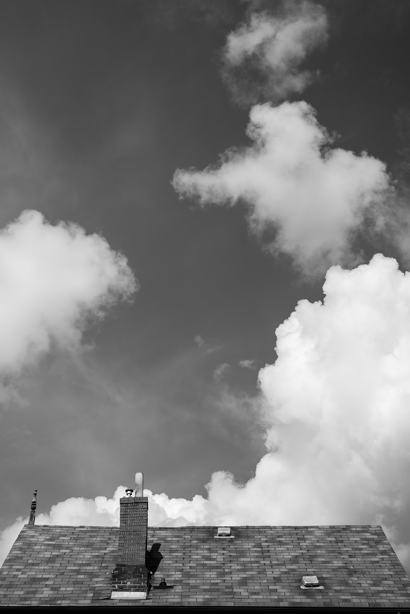 Black-and-white photo of rooftops with a chimney and vents under a sky with large, fluffy clouds.
