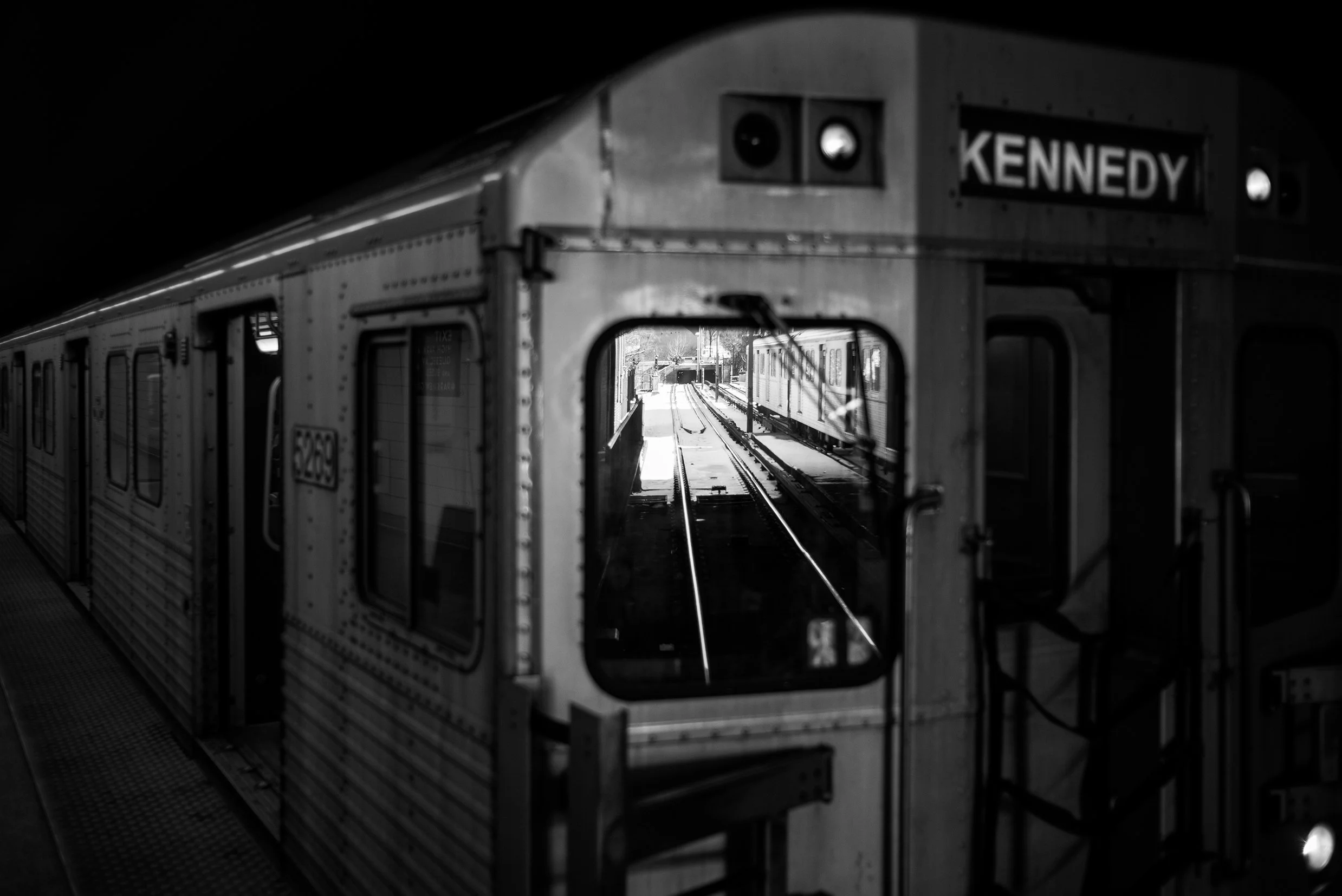 Black and white photo of a subway train at a station, with a sign reading 'Kennedy' on the front of the train.”}