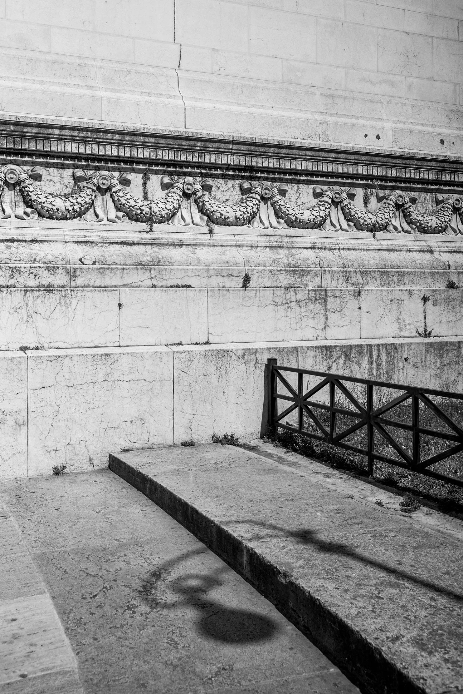 Black and white photo of an old stone wall with decorative sculpted garland motif, a concrete sidewalk, and a small black metal railing, with a shadow of a streetlamp on the ground.