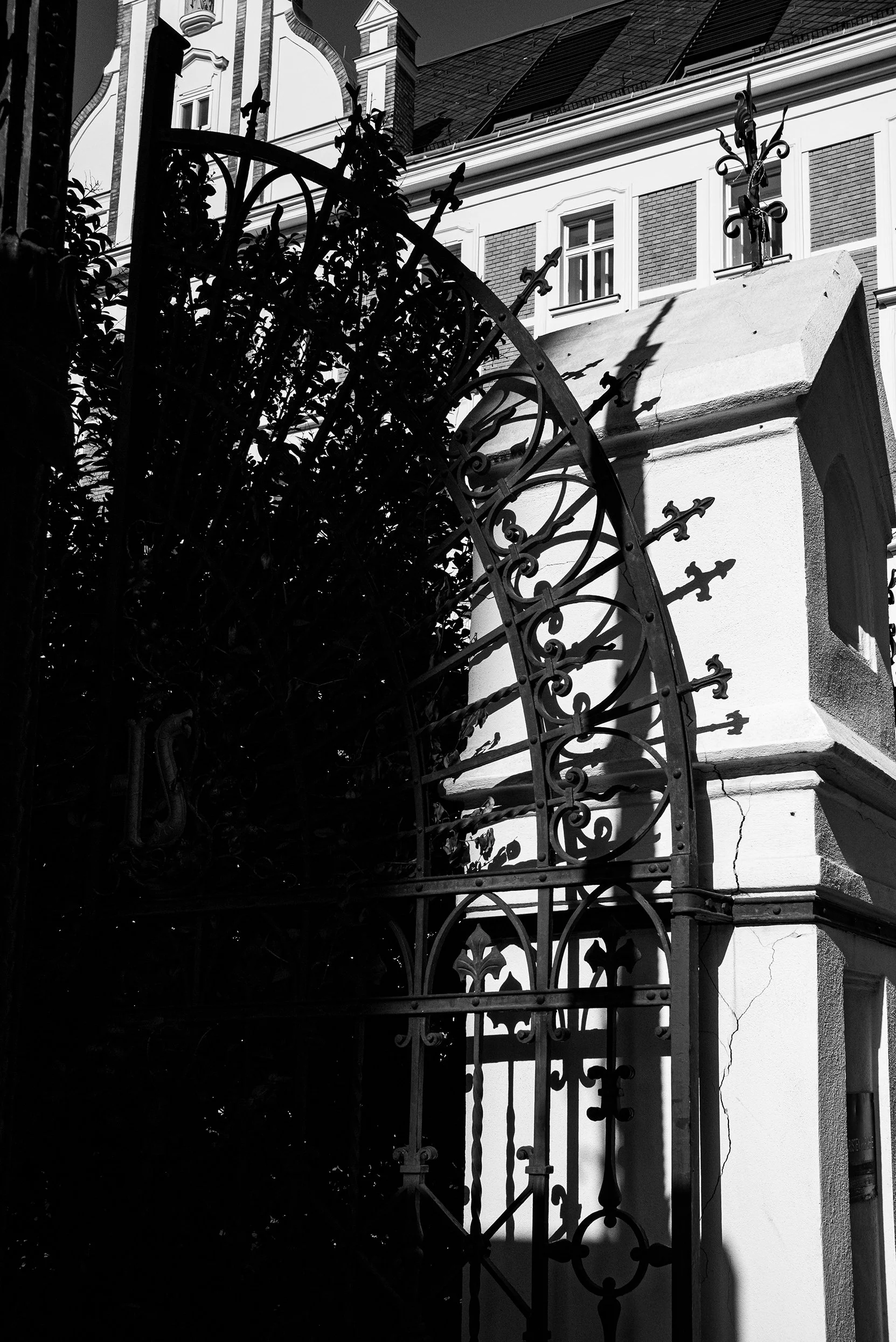 Black and white photo of an ornate metal gate attached to a stone archway, with a building featuring several windows in the background.