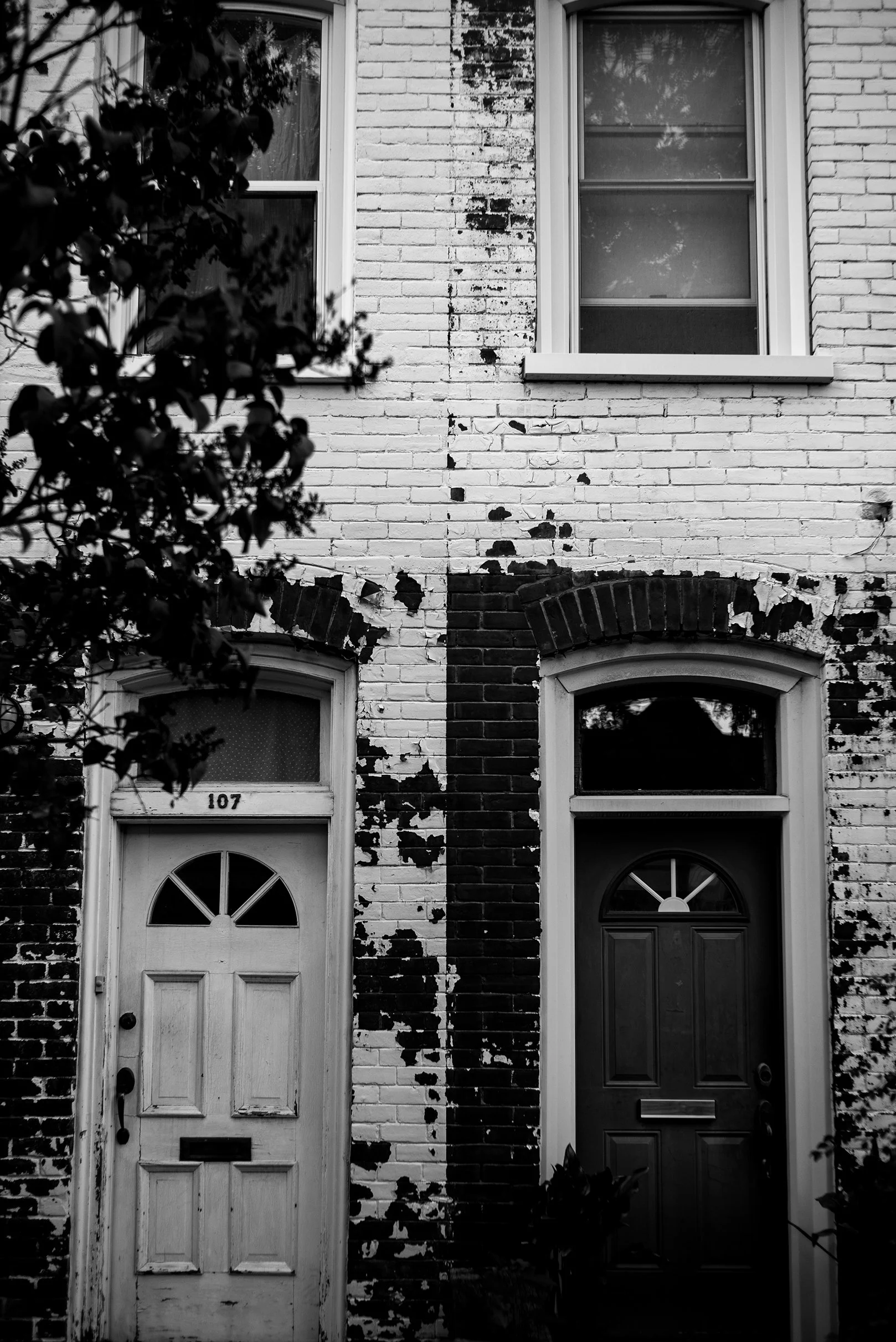 Black and white photograph of two adjacent front doors on a brick building, with peeling paint and four windows above, partially obscured by a tree on the left.