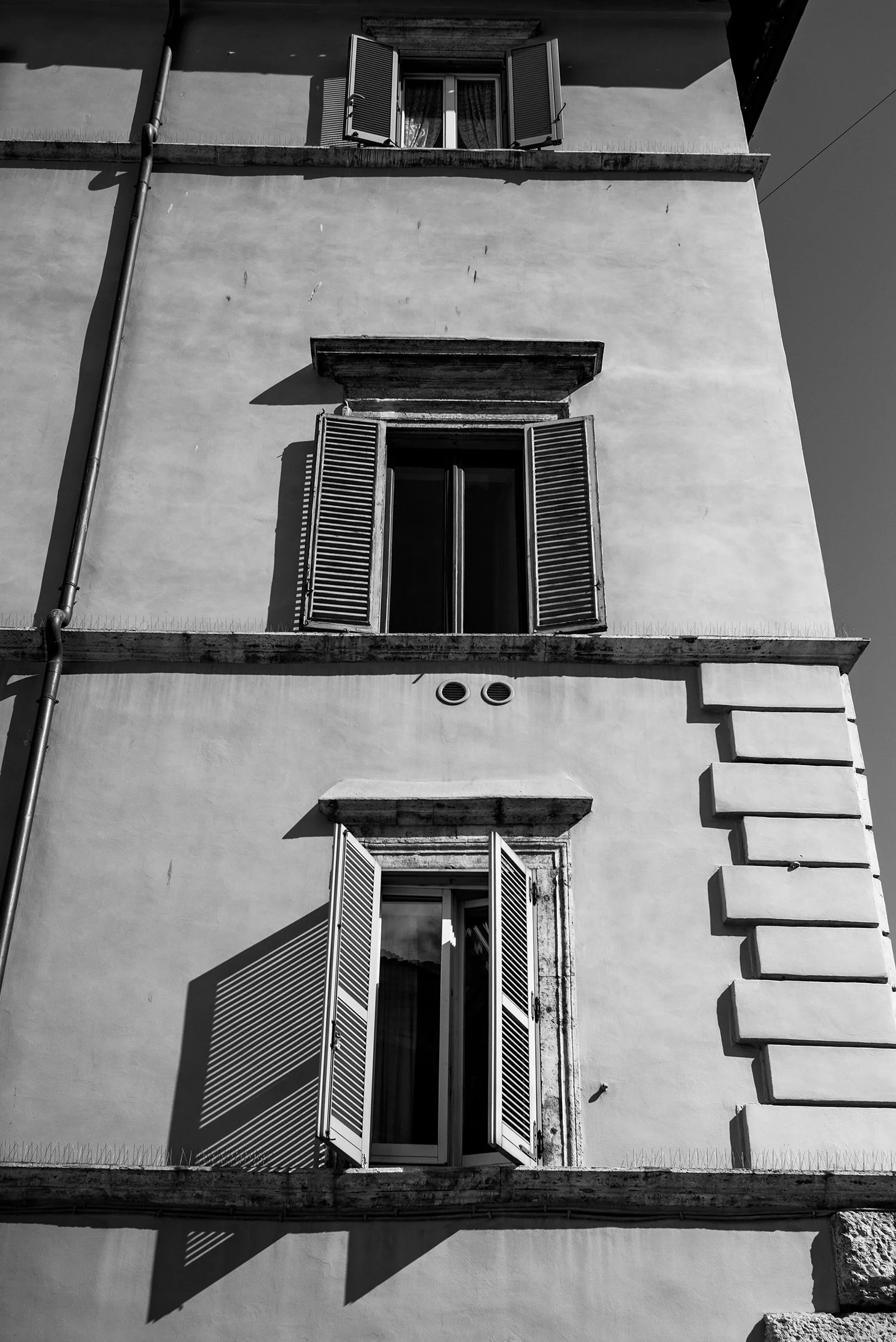 Black and white photo of a building with three windows, each with open shutters. Shadows cast by the shutters create patterns on the facade. There is a drainpipe on the left side and decorative stonework around the windows.