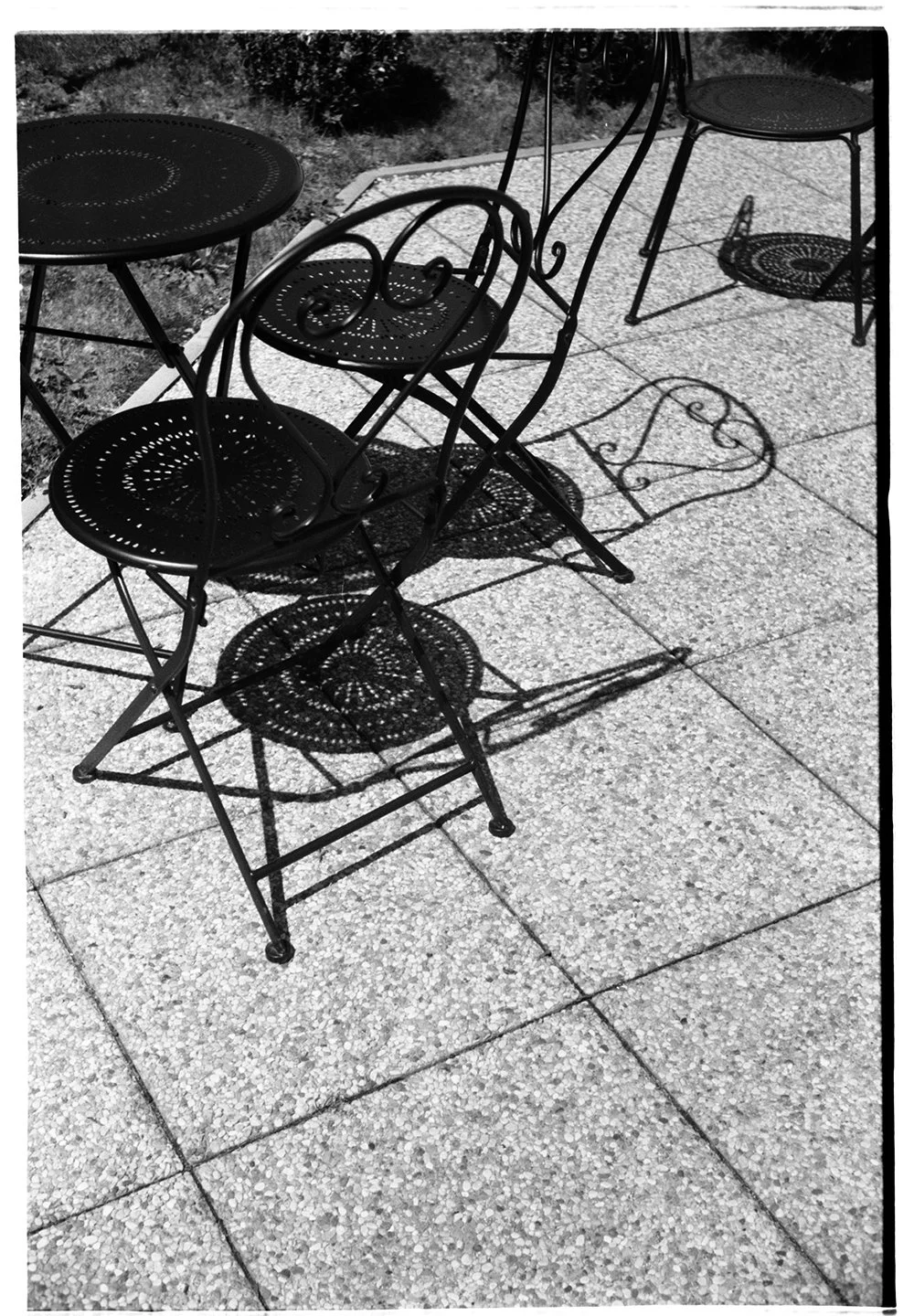 Black wrought iron patio table with two matching chairs on a concrete porch, with shadows cast on the ground.