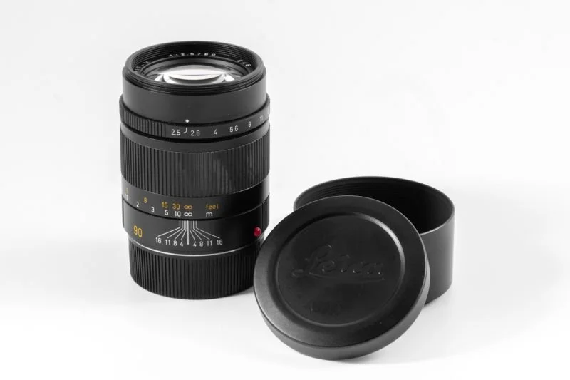 A Lecia camera lens with a black lens cap next to it, both placed on a white background.