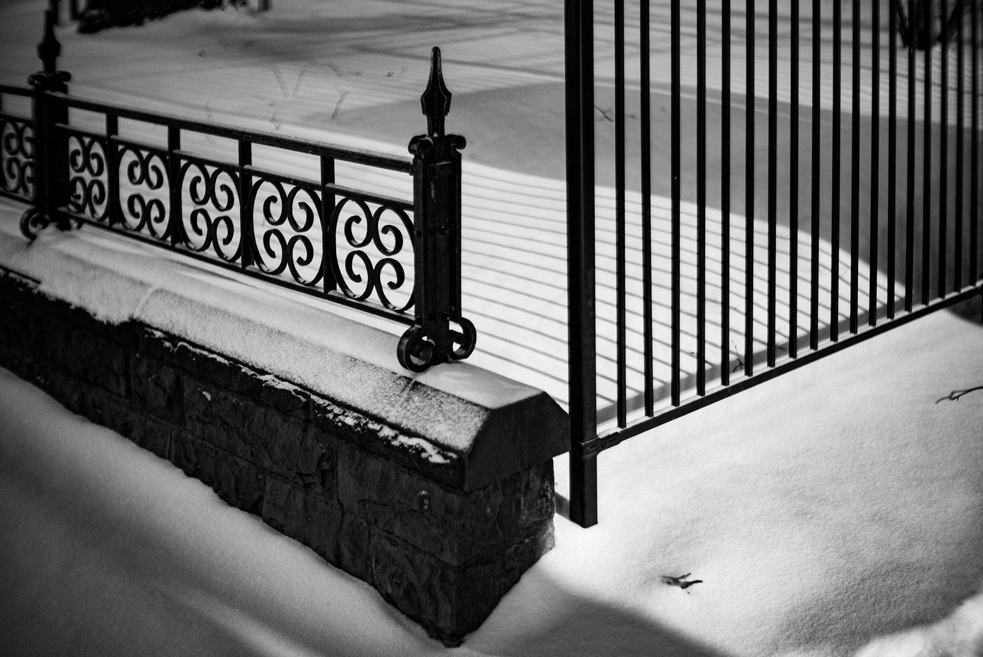 Black and white photo of a snow-covered wrought iron fence and stone base, with snow on the ground and a few footprints.
