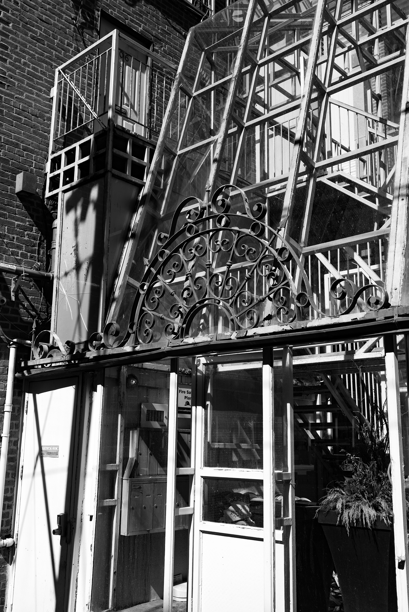 Black and white photo of an outdoor fire escape with ornate metalwork, glass panels, and stairs on the side of a brick building.