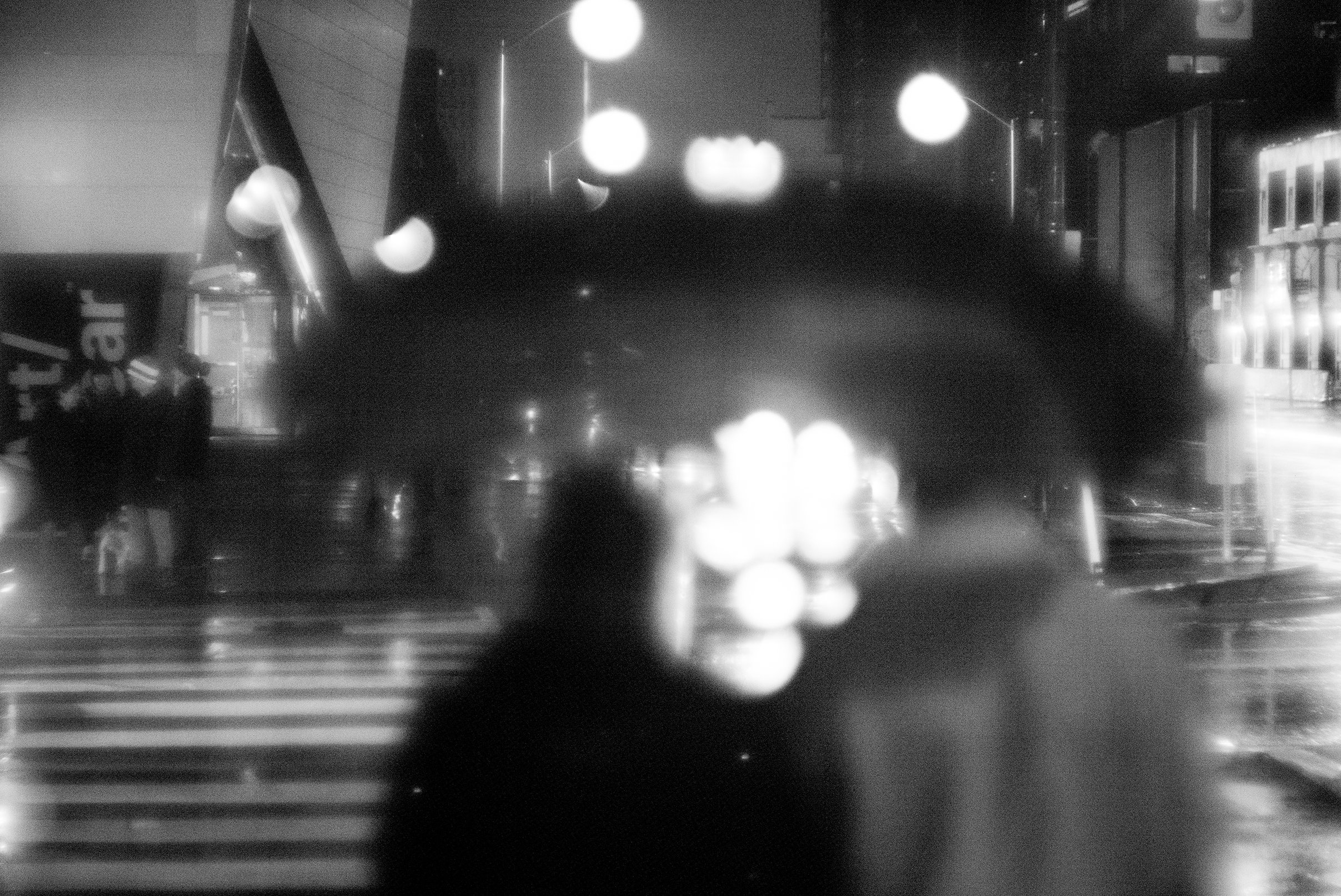 A black and white photo taken at night of a city street with blurred lights. The photo is taken through a rain-covered window, showing an urban scene with a blurred silhouette of a person and a crosswalk.