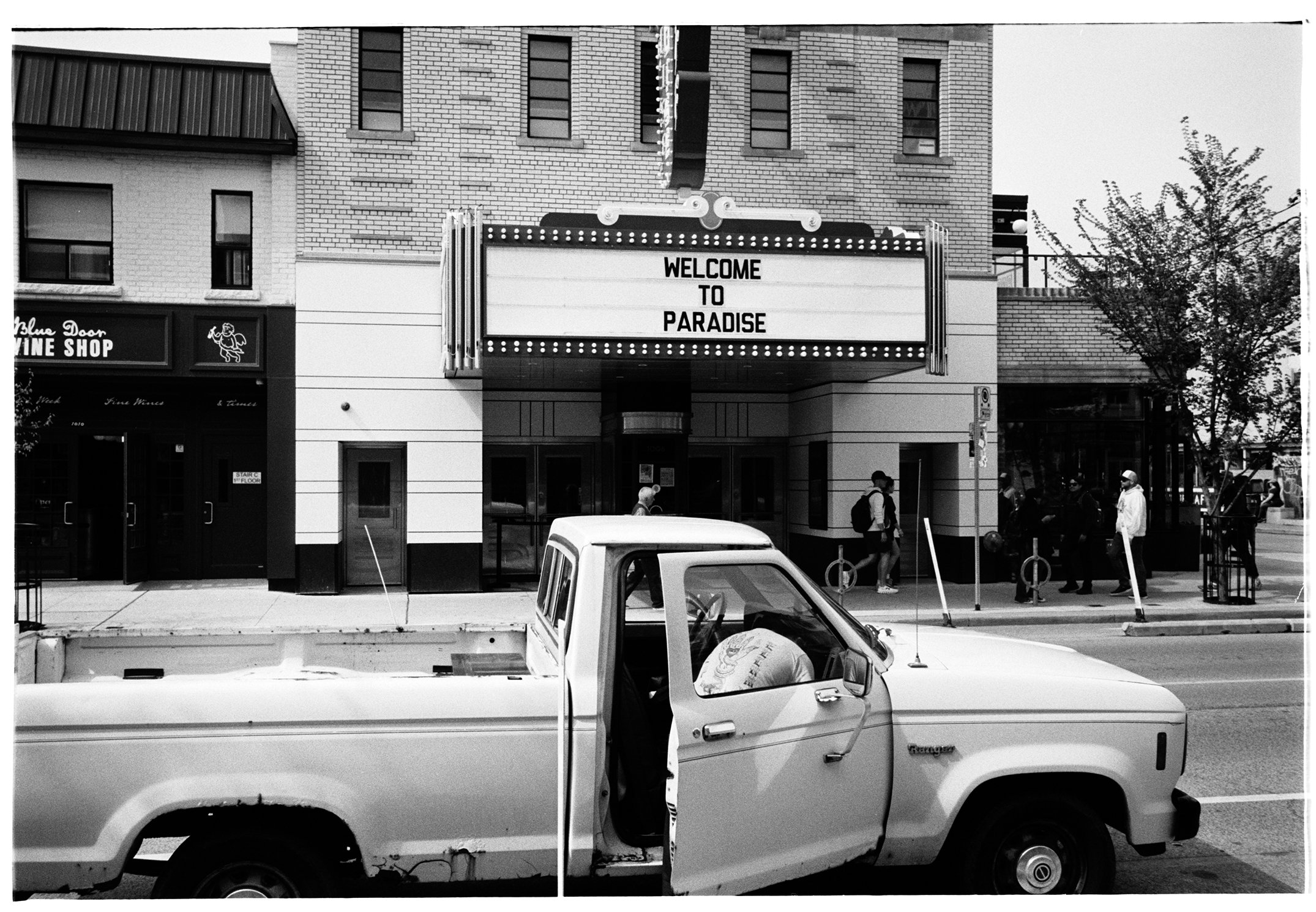 Street scene in front of a theater with a sign that says "Welcome to Paradise." A pickup truck with an open door is in the foreground, and several pedestrians are walking on the sidewalk.