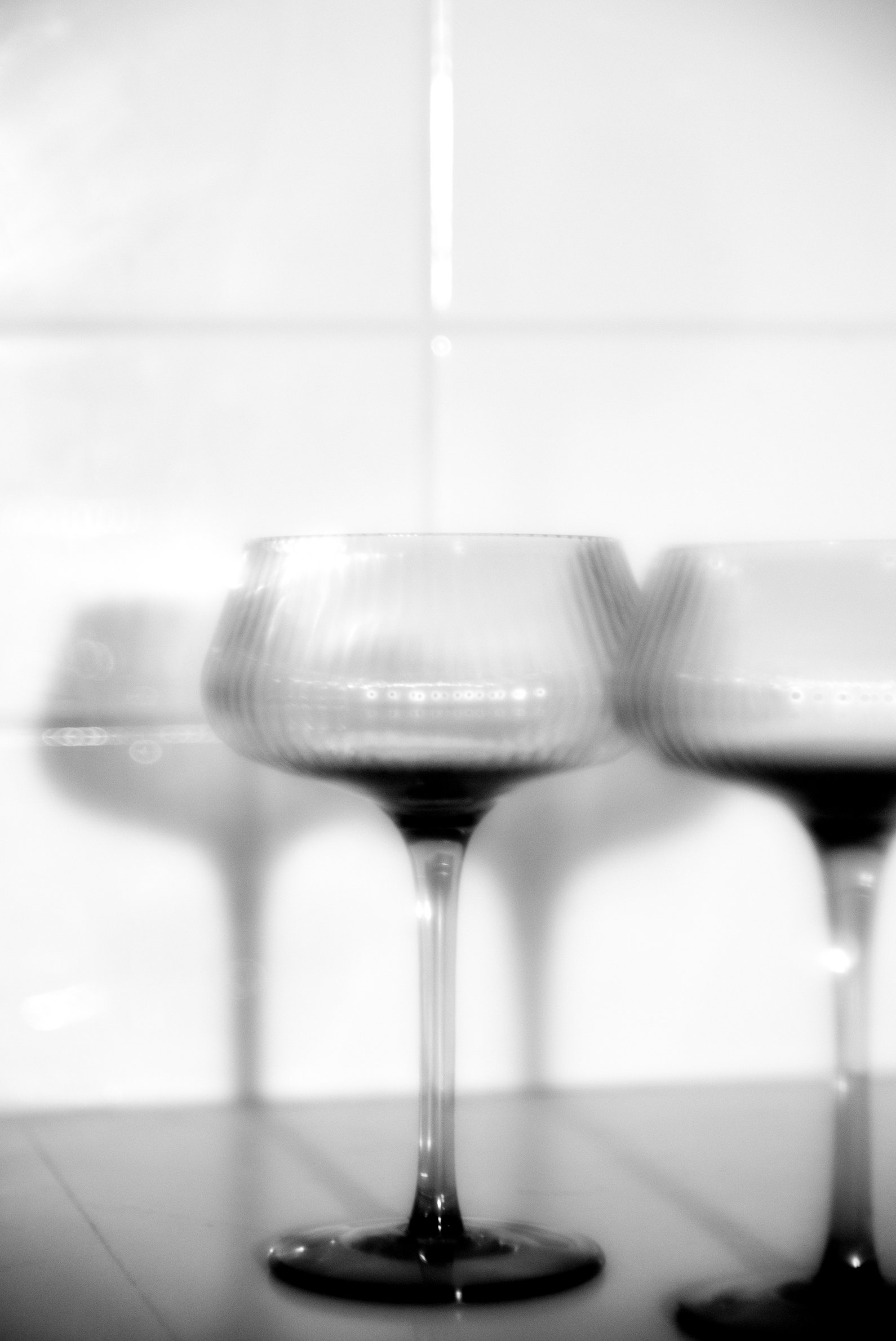Black and white image of three wine glasses, with one in focus in the center, against a tiled wall background.