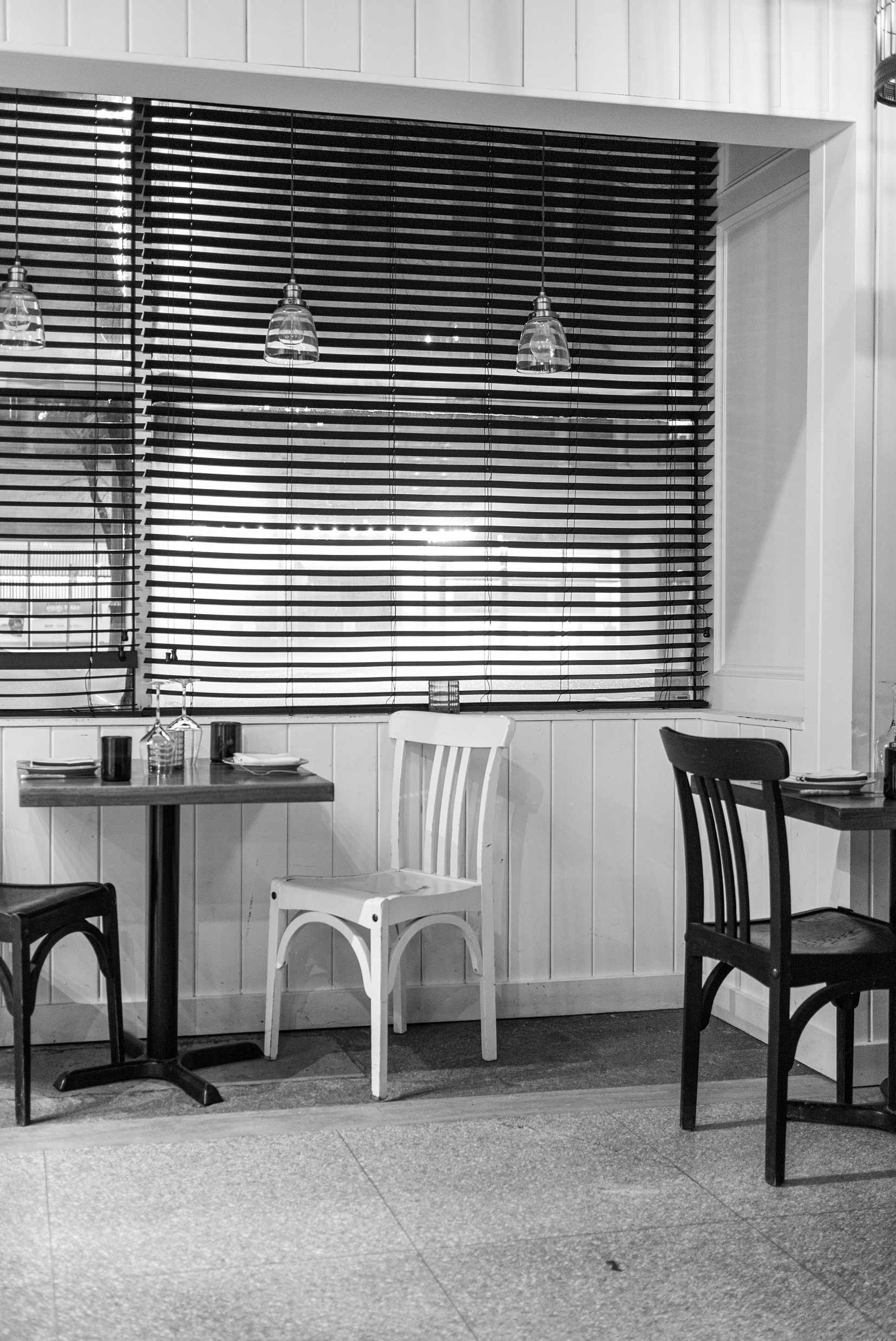 Interior of a restaurant with wooden panel walls, window with blinds, hanging pendant lights, and empty tables with chairs.