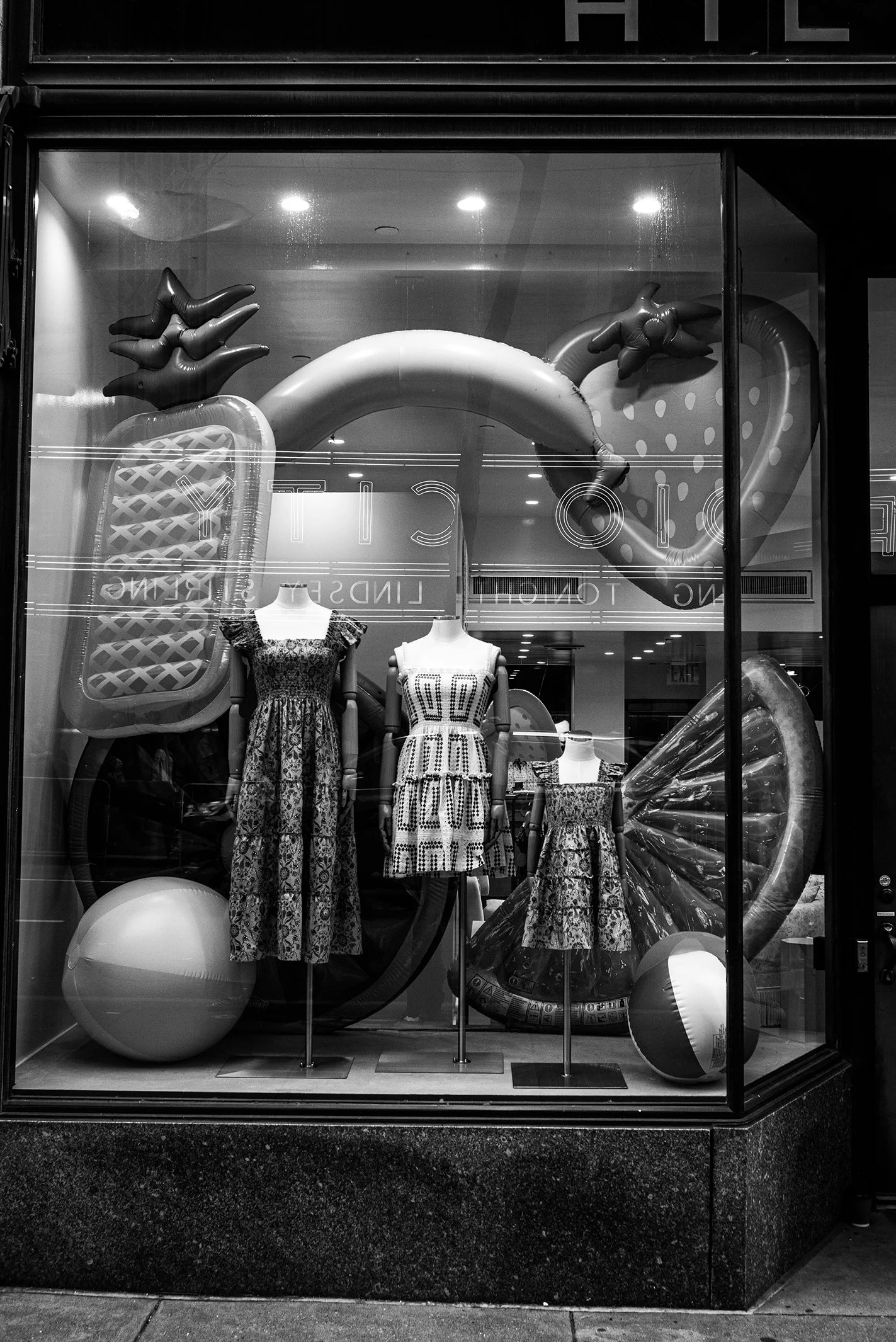 Display window with mannequins wearing patterned dresses and inflatable summer items, including a pineapple, ice cream, popsicle, and lemon slice, in black and white.