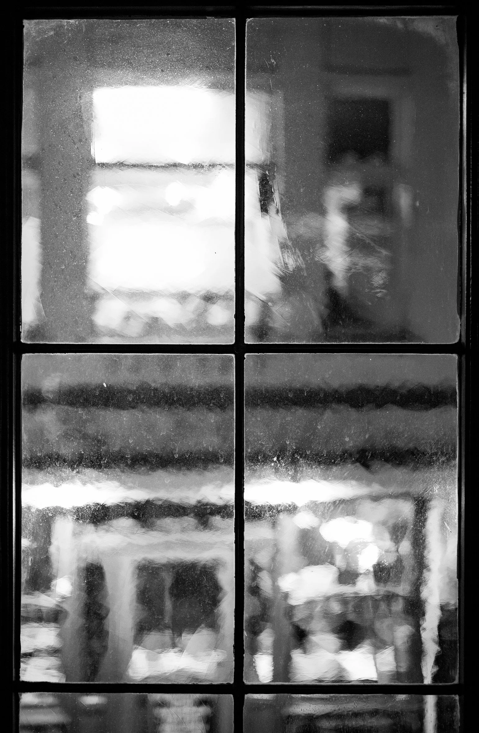 View through a dirty glass window showing a blurred reflection of a person, shelves, and lights inside a room, in black and white.