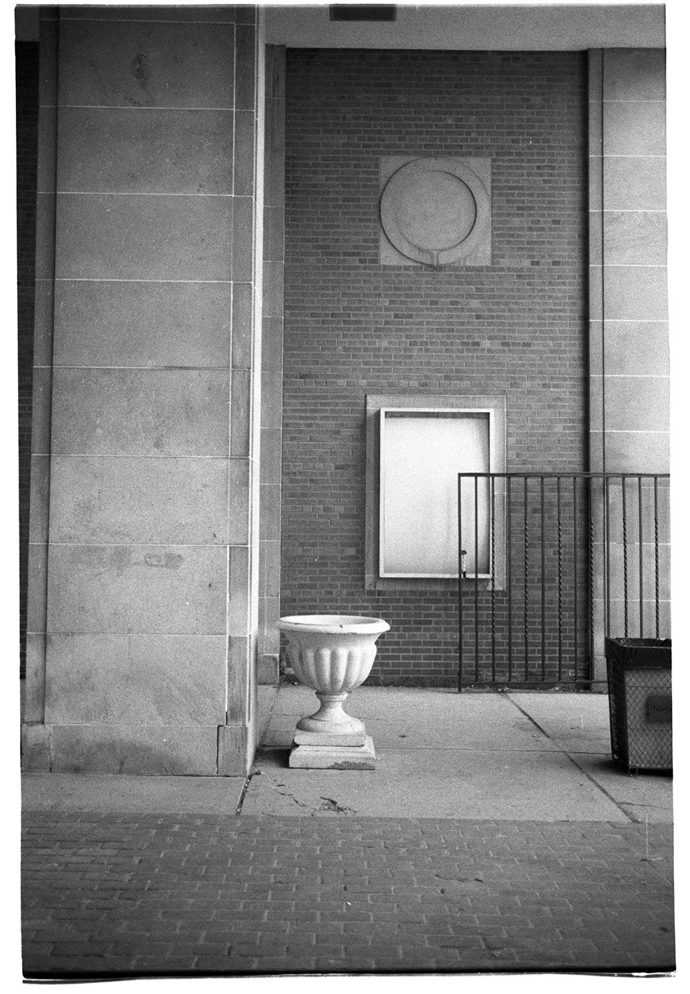 Black and white photo of a sidewalk scene with a brick wall, a decorative planter, a blank framed notice, and a trash bin.