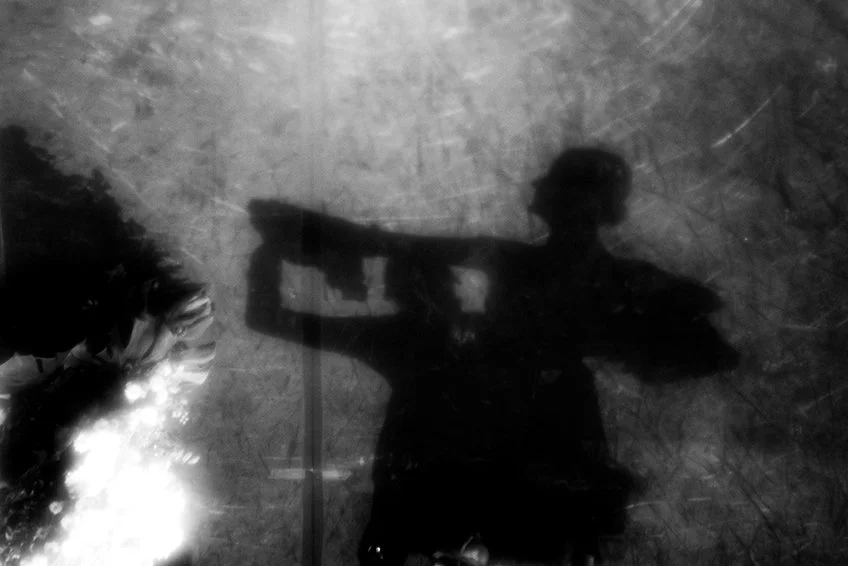 Silhouette of a person taking a photo, reflected on a surface, with a partial view of a flower on the left side.
