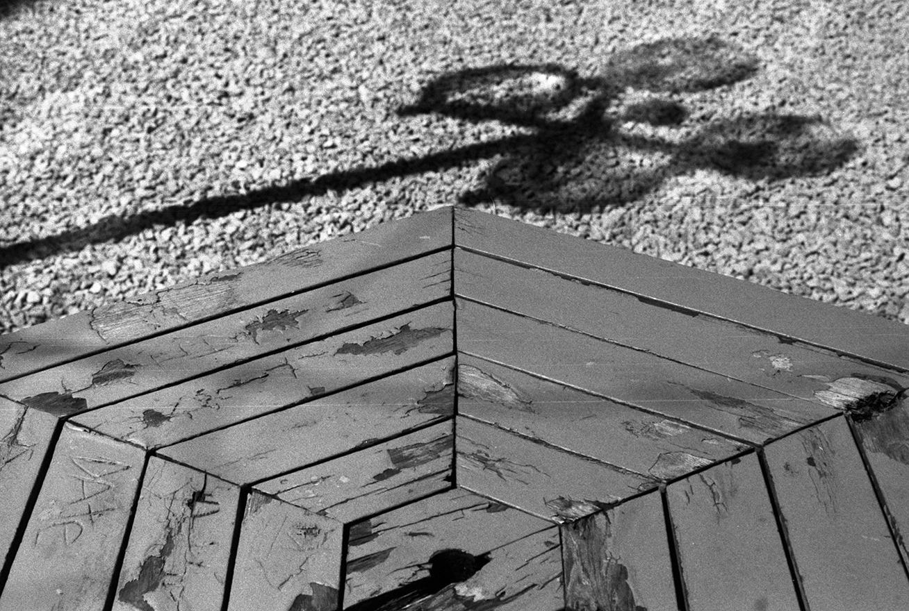 Black and white photo showing a wooden corner with peeling paint, with a small hole near the bottom, and a pair of scissors cast in shadow on a textured gravel ground above.