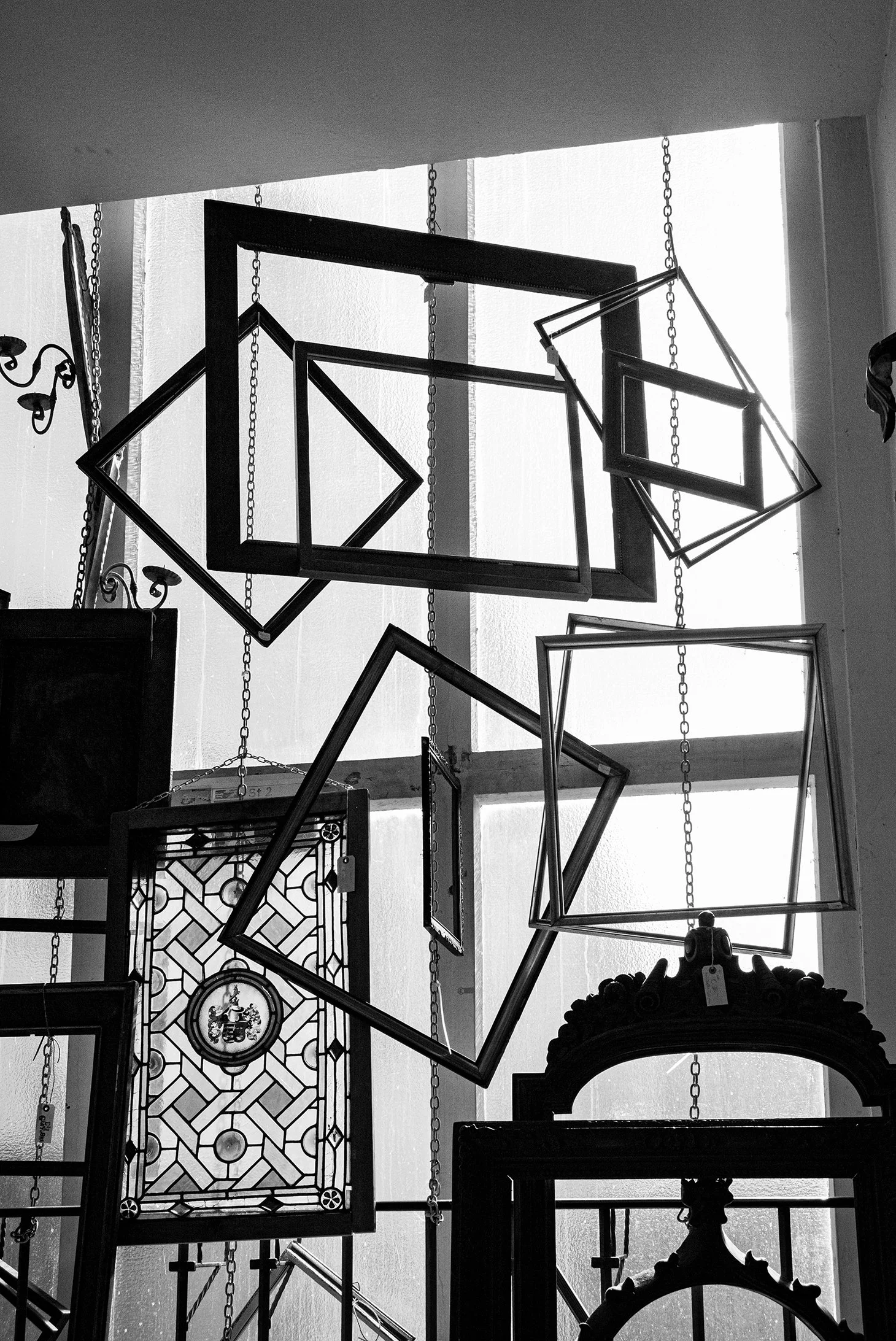 Black and white photo of empty picture frames hanging from chains against a bright window.