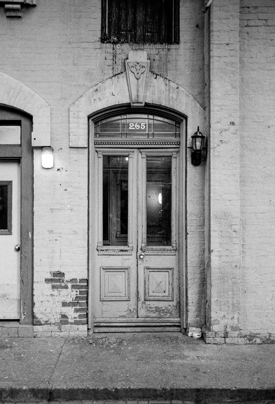 Black and white photo of an old wooden door with glass panels, set in a brick building with the number 265 above it. There is graffiti or peeling paint on the brick wall to the left of the door, a wall-mounted lantern on the right, and a small window above.