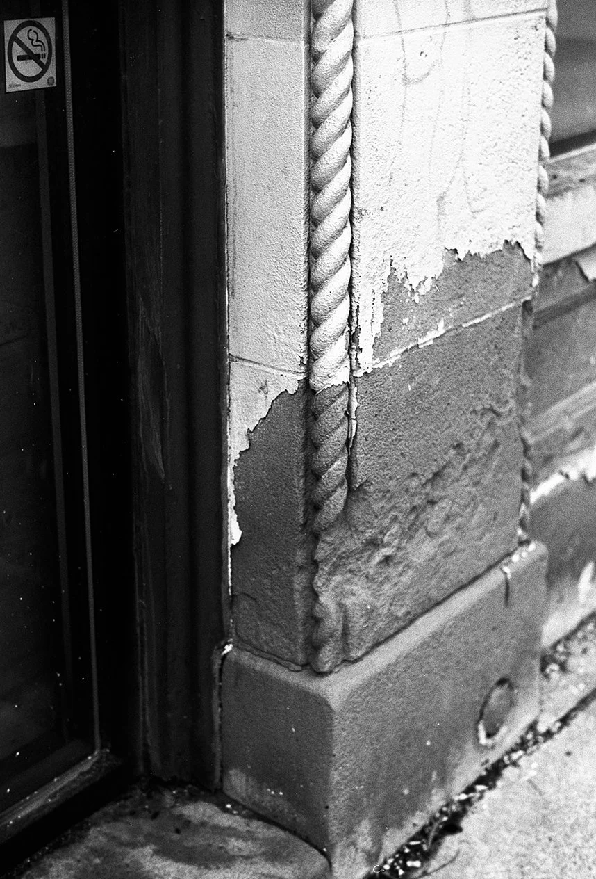 Close-up of a corner of a brick building with a rusted metal rope or cable attached to the wall, and a small sign indicating a no-smoking area next to a window.