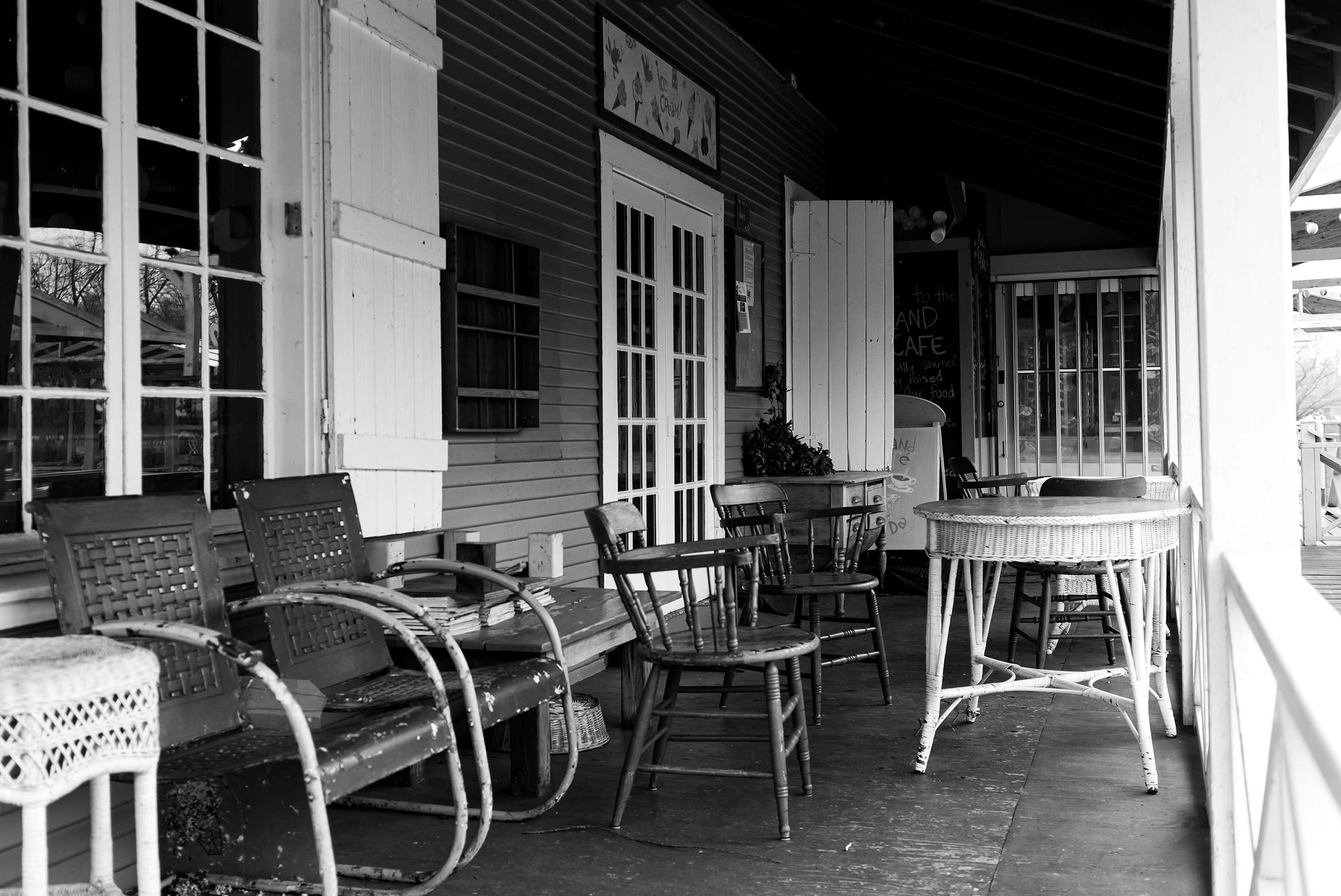 Black and white photo of a rustic cafe porch with wooden chairs and tables, some with peeling paint, outside a building with open shutters and a sign.